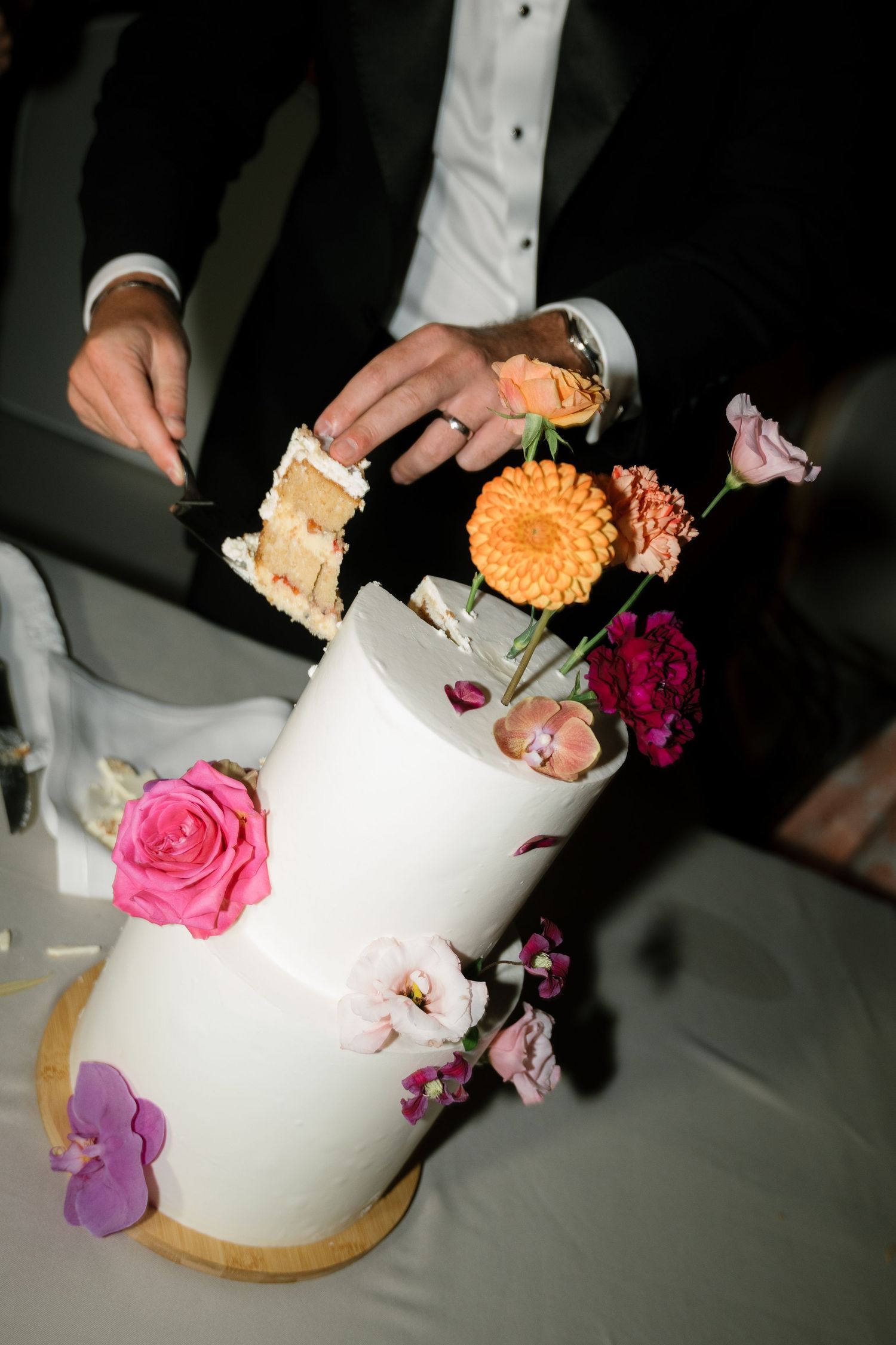 A white wedding cake decorated with pink, purple and orange flowers being cut into slices.