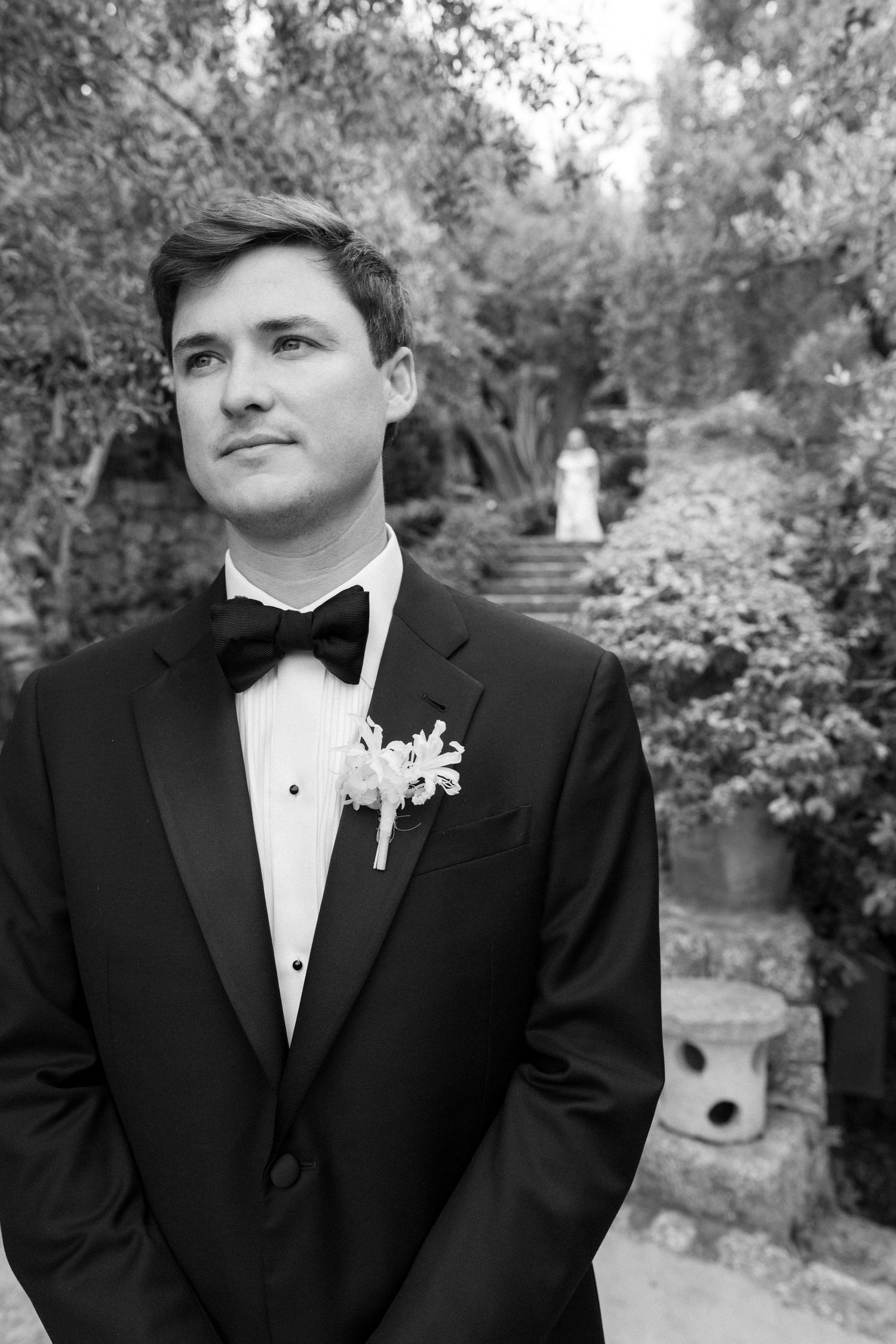 A person in a black tuxedo and bowtie poses outdoors in a garden setting during a formal event.