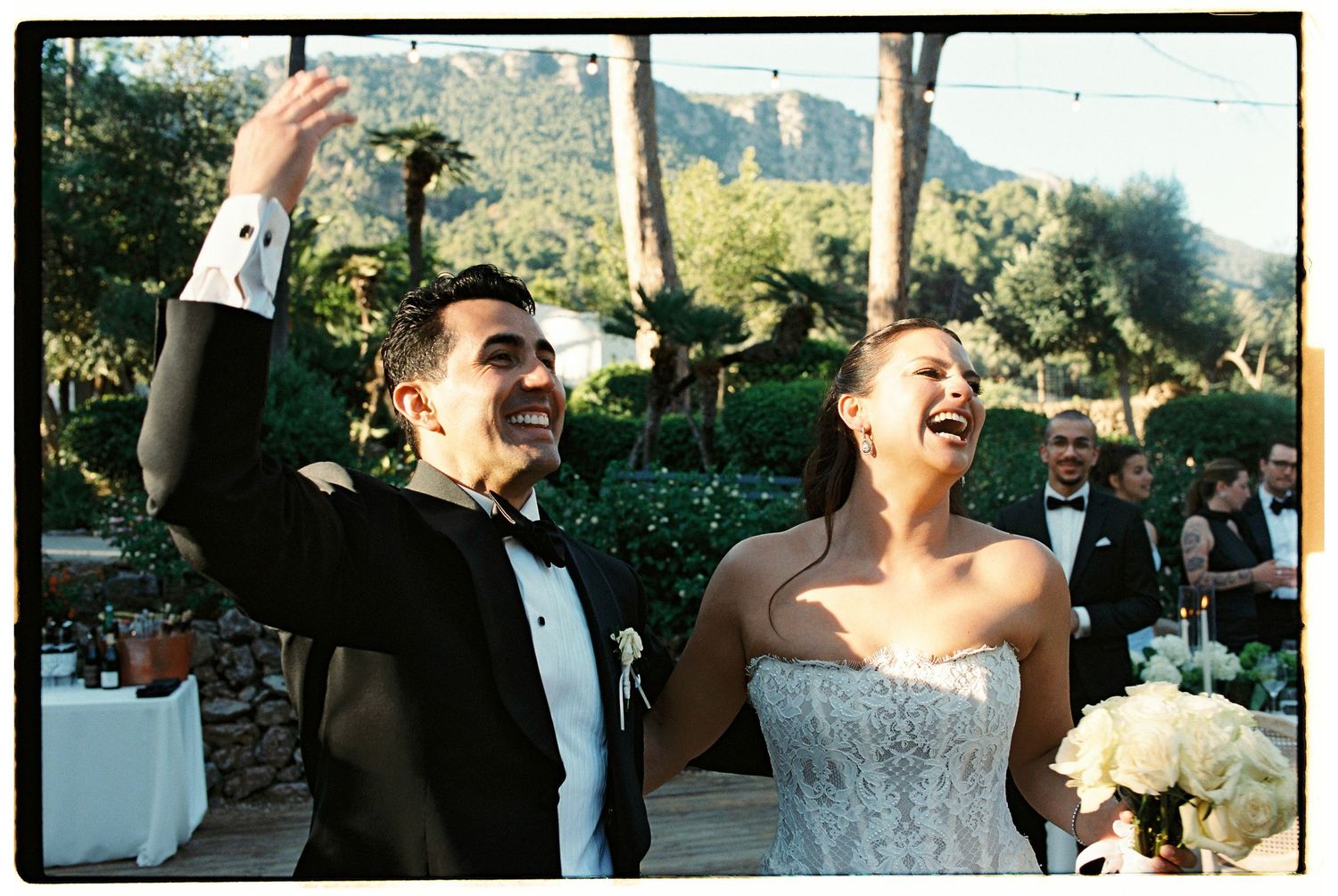 Joyful bride and groom laughing and celebrating at outdoor wedding reception with lush green mountains in background.