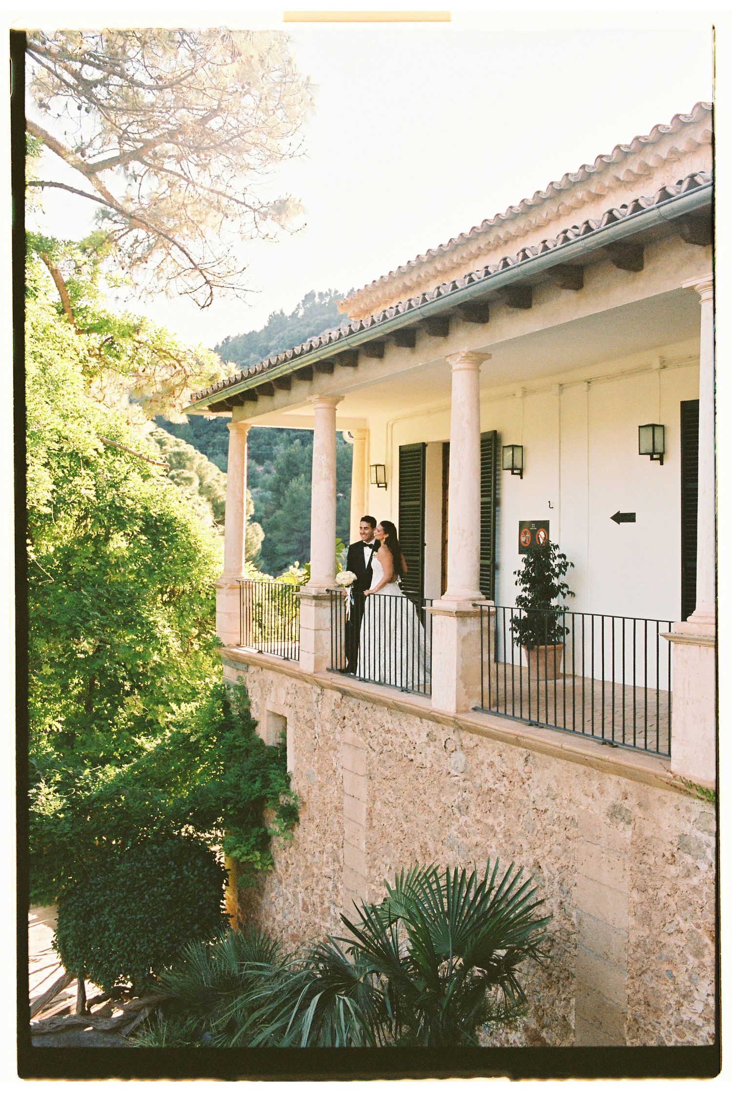 Bride and groom stand on elegant villa balcony surrounded by lush greenery during outdoor wedding celebration.