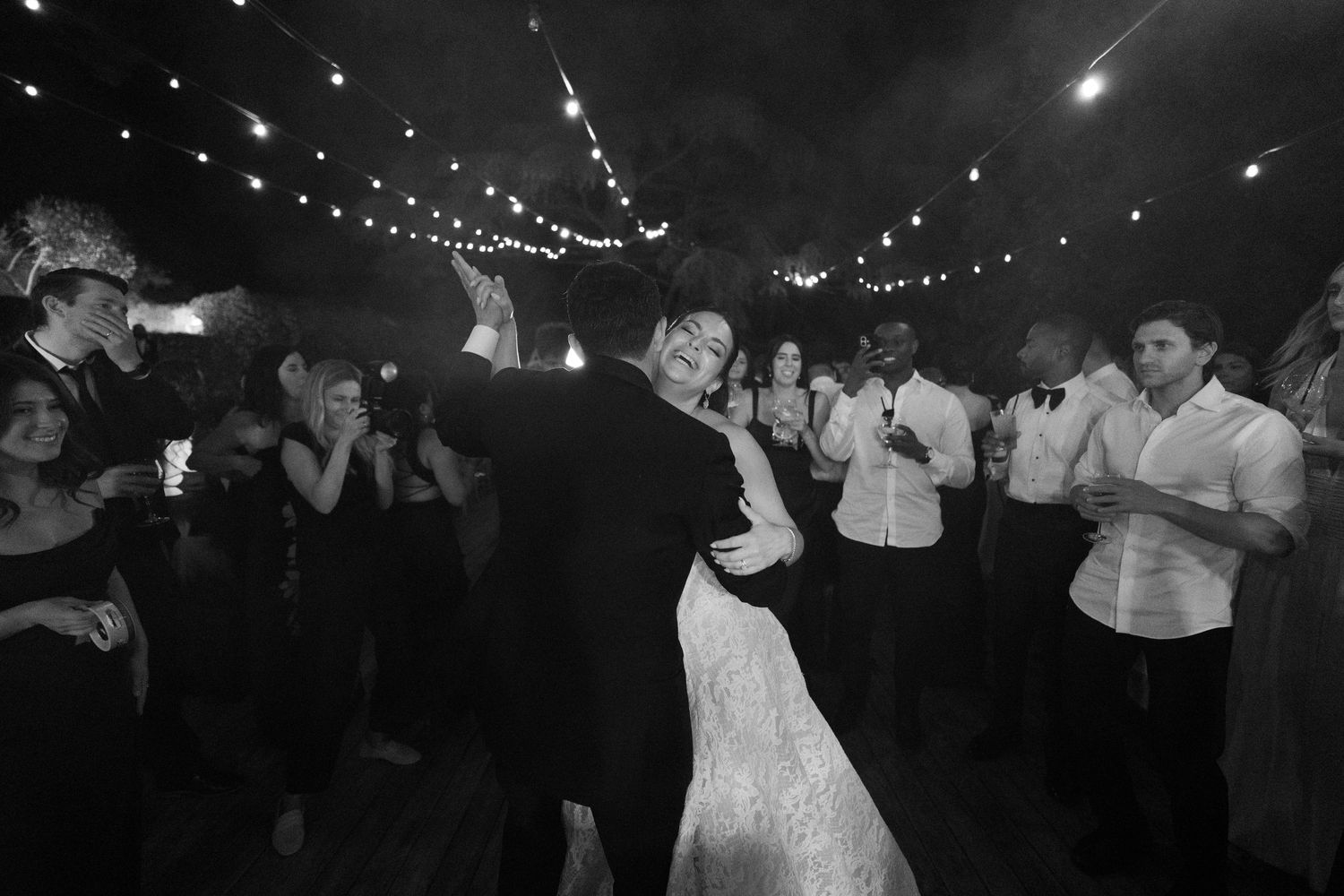 Black and white photo of a bride and groom sharing their first dance surrounded by guests under string lights.