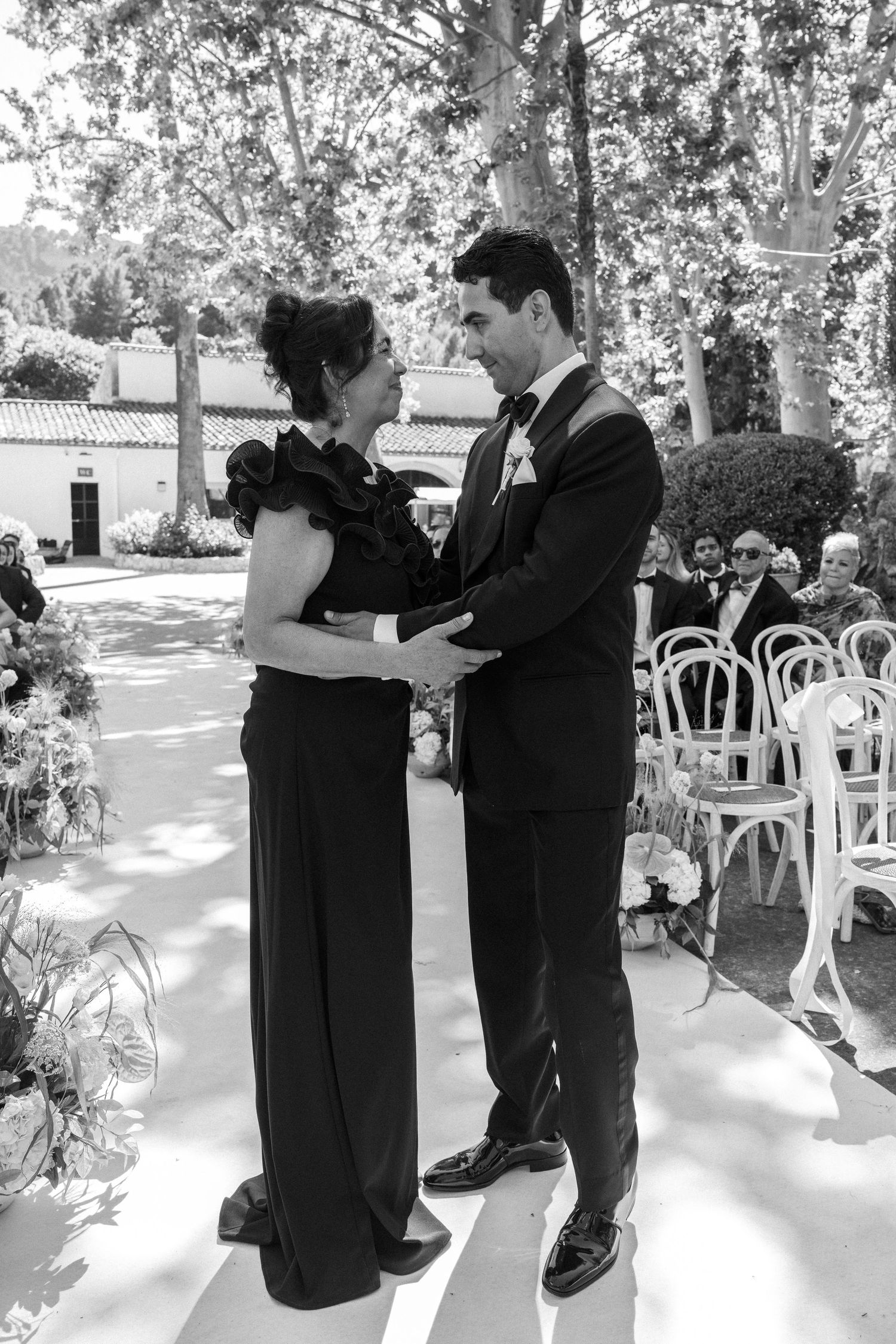 Black and white photo of groom and mother sharing a dance at outdoor wedding ceremony with white chairs.