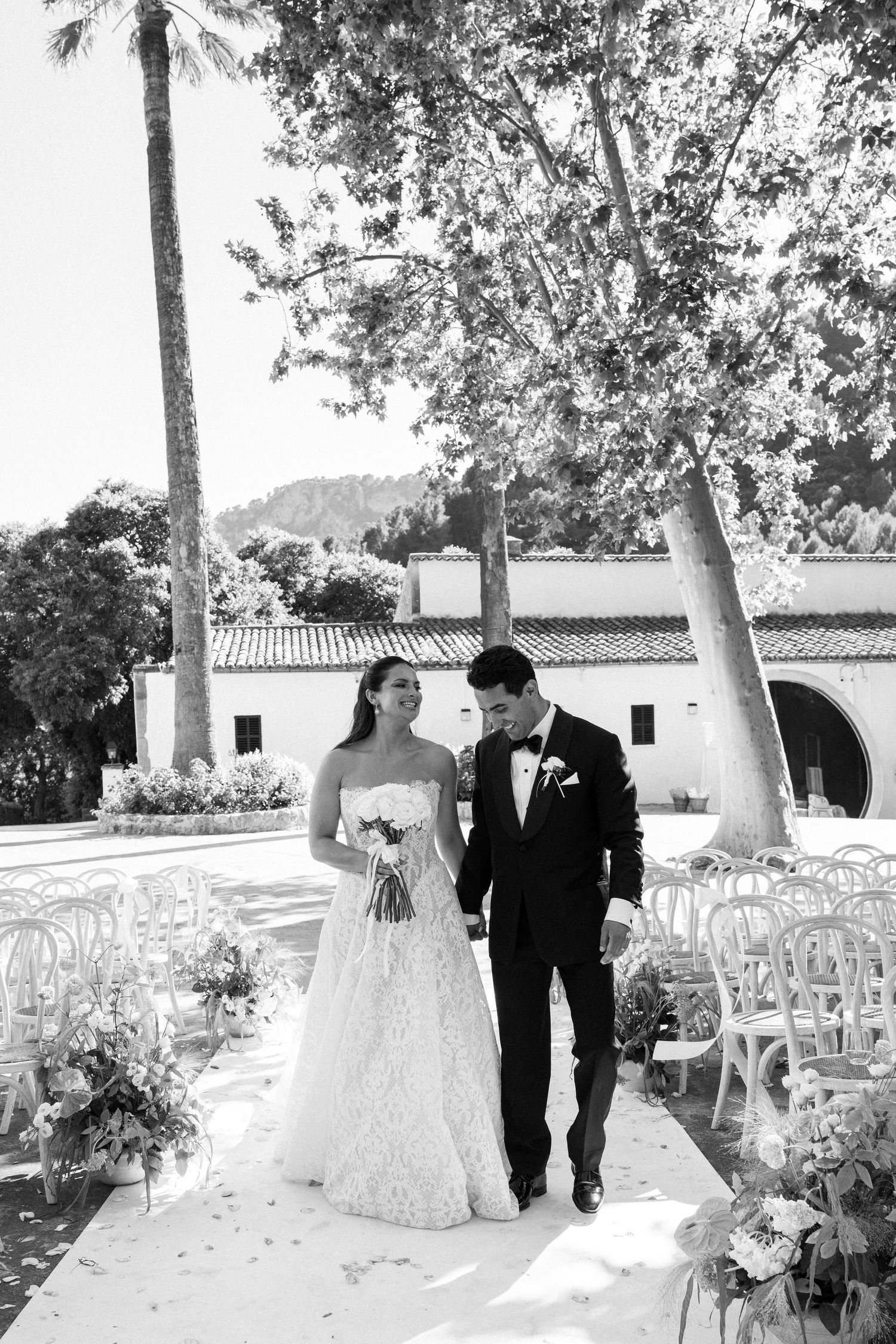 Black and white photo of bride and groom walking down outdoor wedding aisle lined with white chairs and floral arrangements.