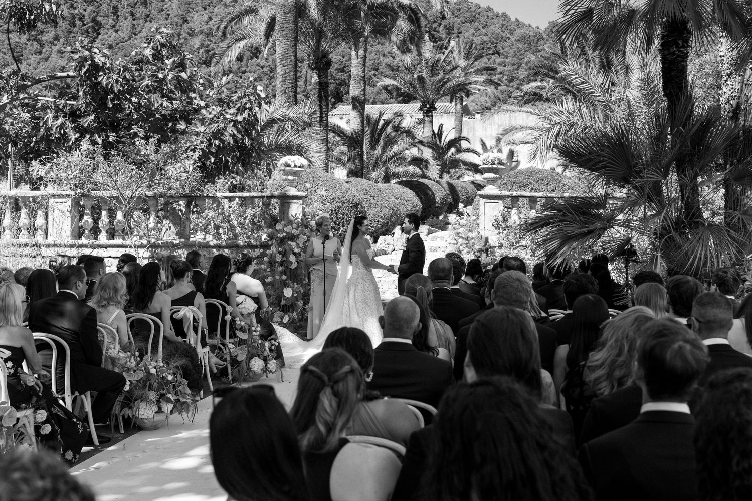 Black and white outdoor wedding ceremony with bride and groom at the altar, surrounded by guests and lush palm trees.