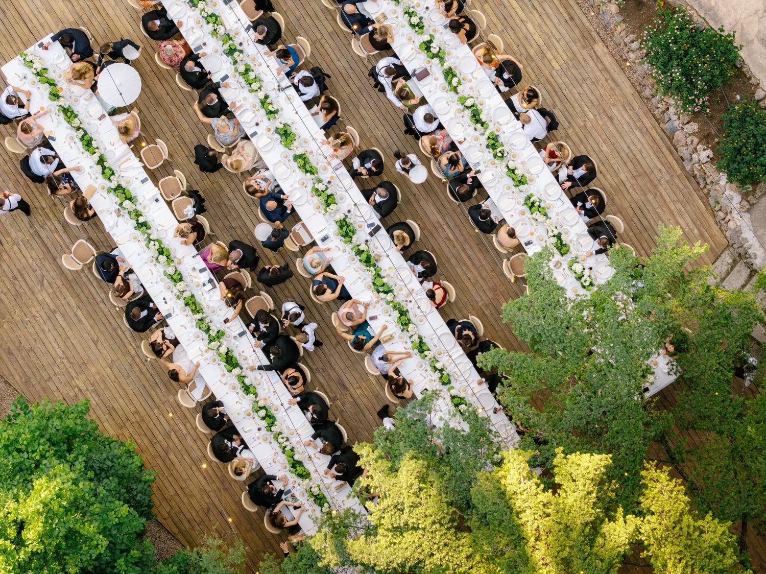 Aerial view of elegant outdoor wedding reception with long white tables, greenery centerpieces, and guests seated on wooden deck.