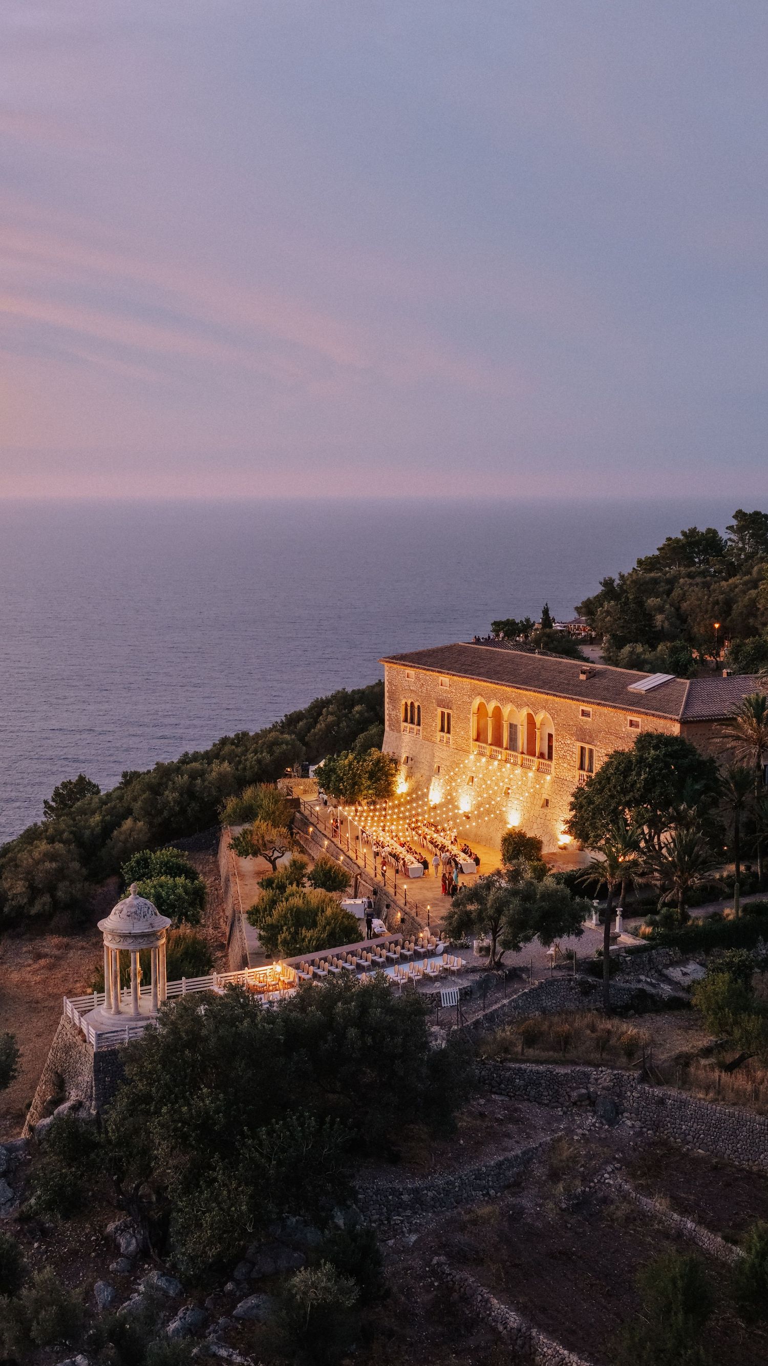 Aerial view of an illuminated cliffside villa Son Marroig with arched architecture surrounded by trees overlooking the Mediterranean Sea.
