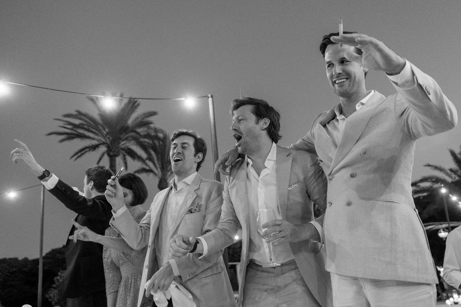 Group of wedding guests in light suits celebrate under string lights at outdoor reception.