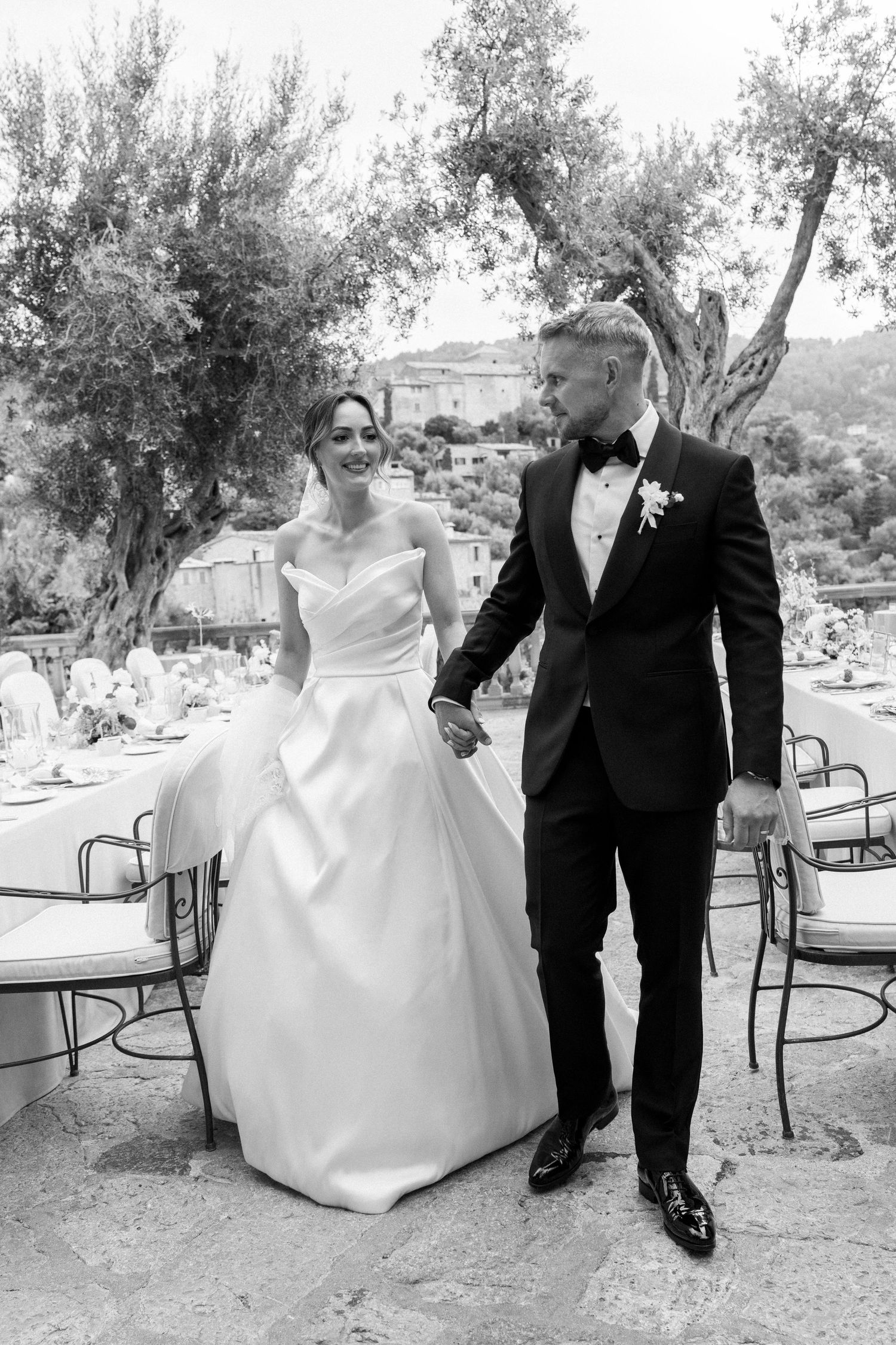 Wedding couple walking together on a rustic outdoor path surrounded by olive trees.