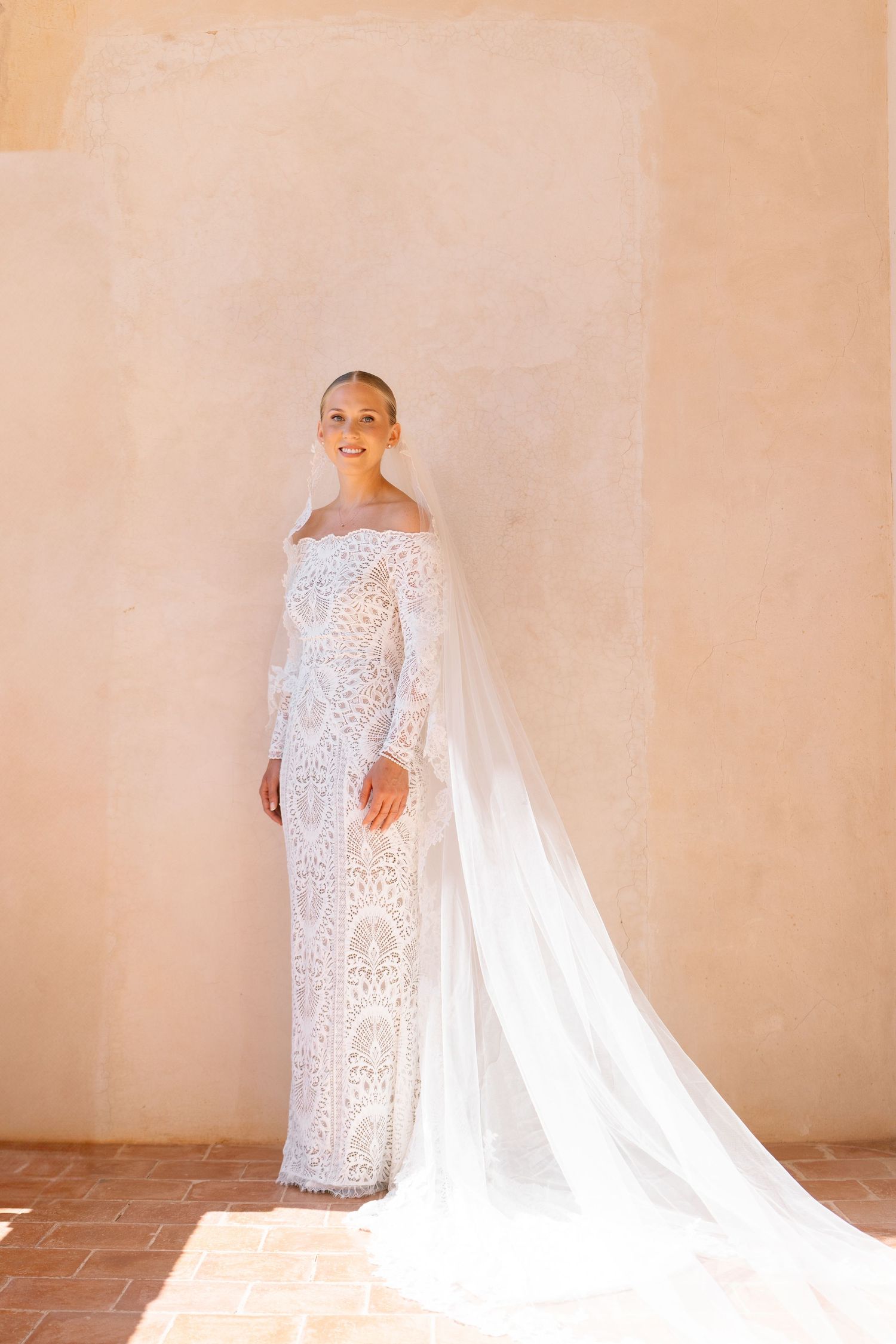 Elegant lace wedding dress with long sleeves and flowing cathedral veil photographed in warm natural light.