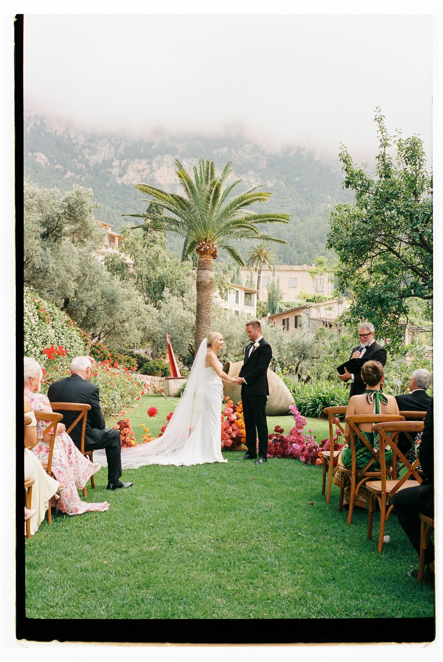 Outdoor wedding ceremony in a lush garden setting with palm trees and mountain views in the background.