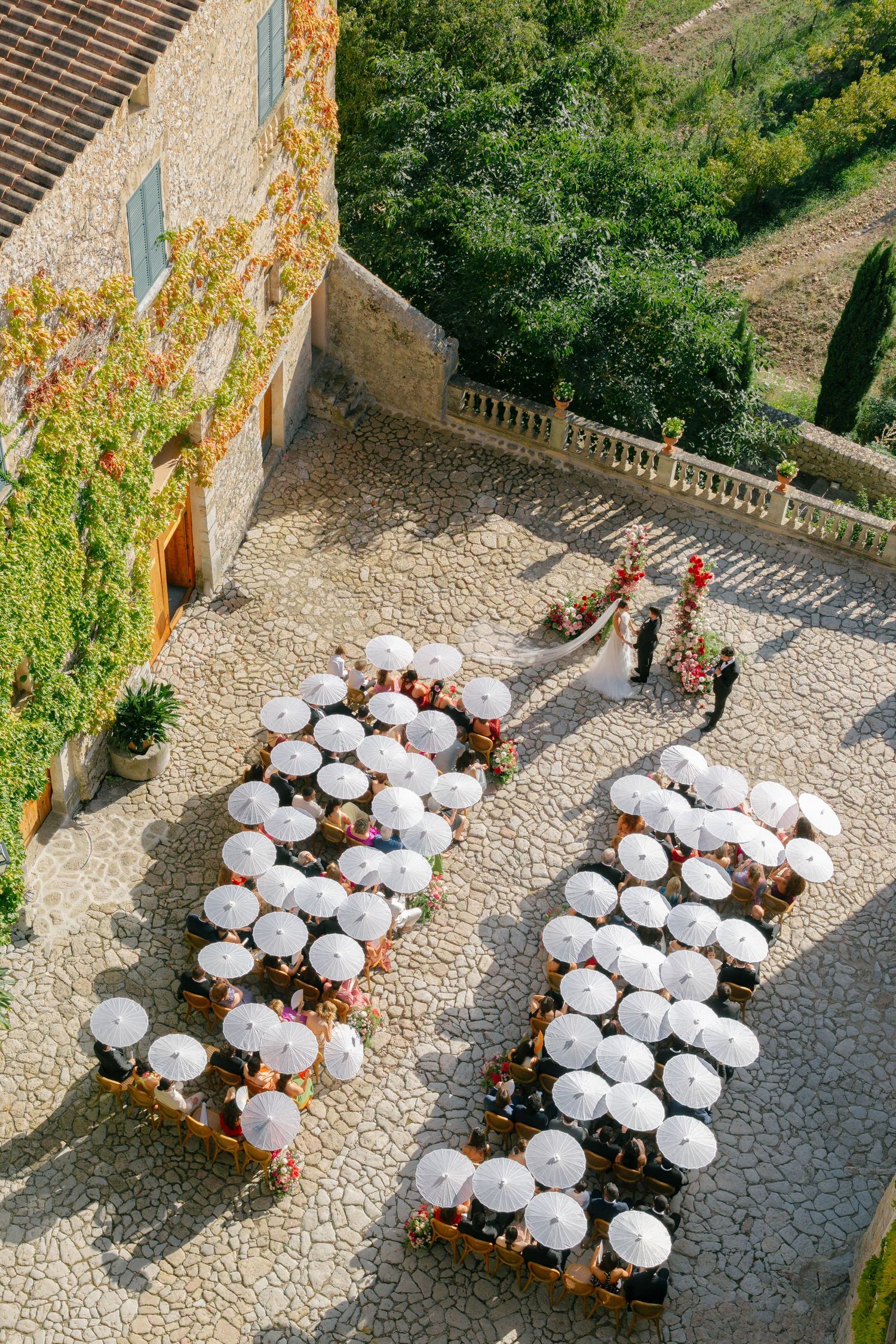 Aerial view of wedding guests with white umbrellas gathered in a cobblestone courtyard on a sunny day.