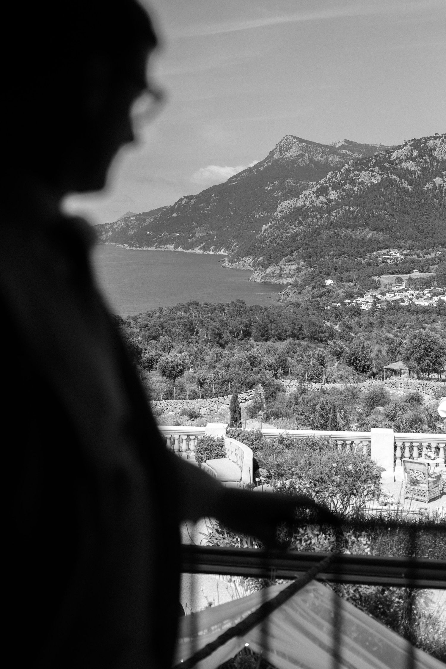 Silhouetted figure gazes at mountainous coastal landscape from balcony.