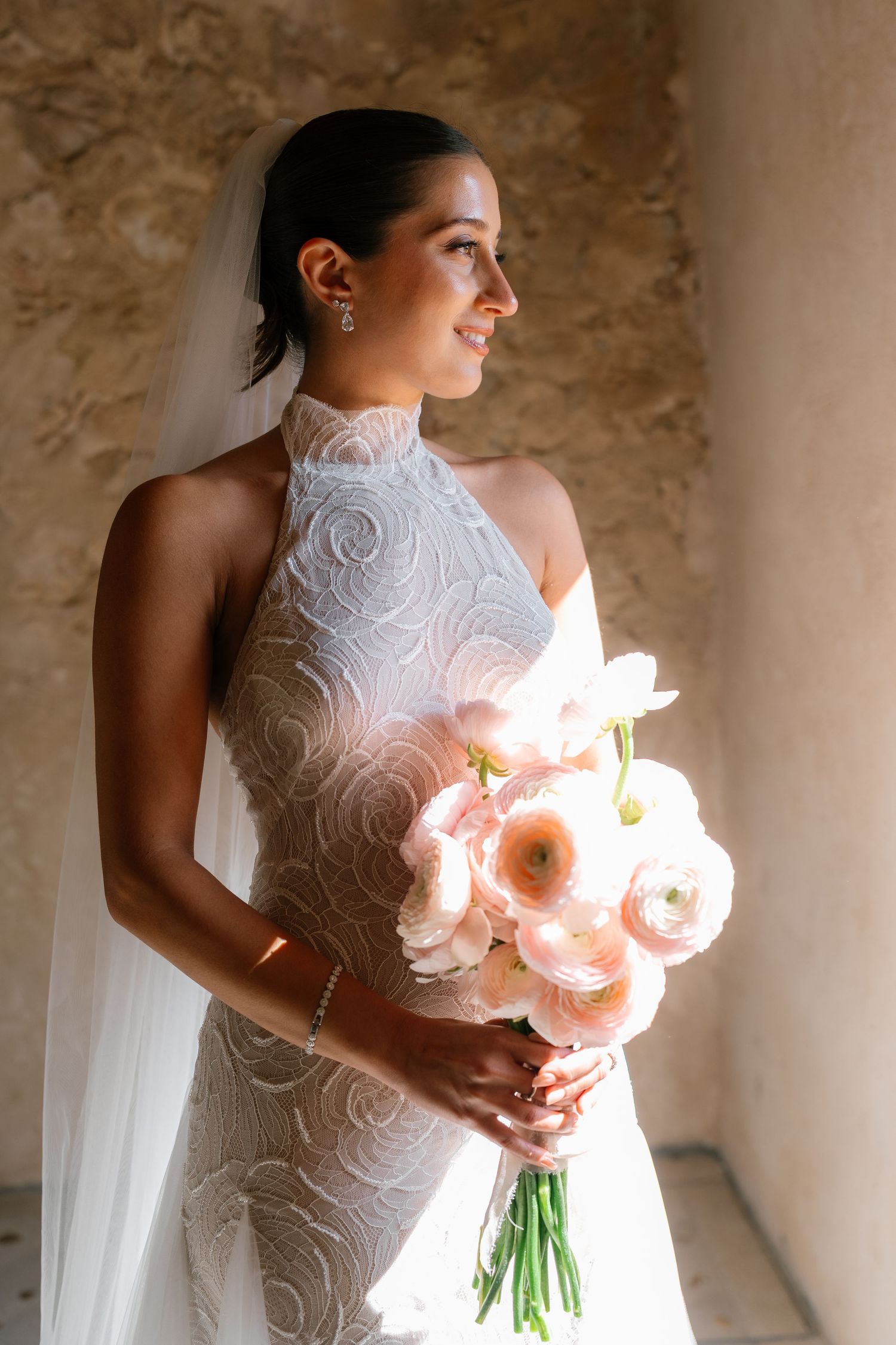 Bride in lace dress holds pink bouquet in sunlit corridor.