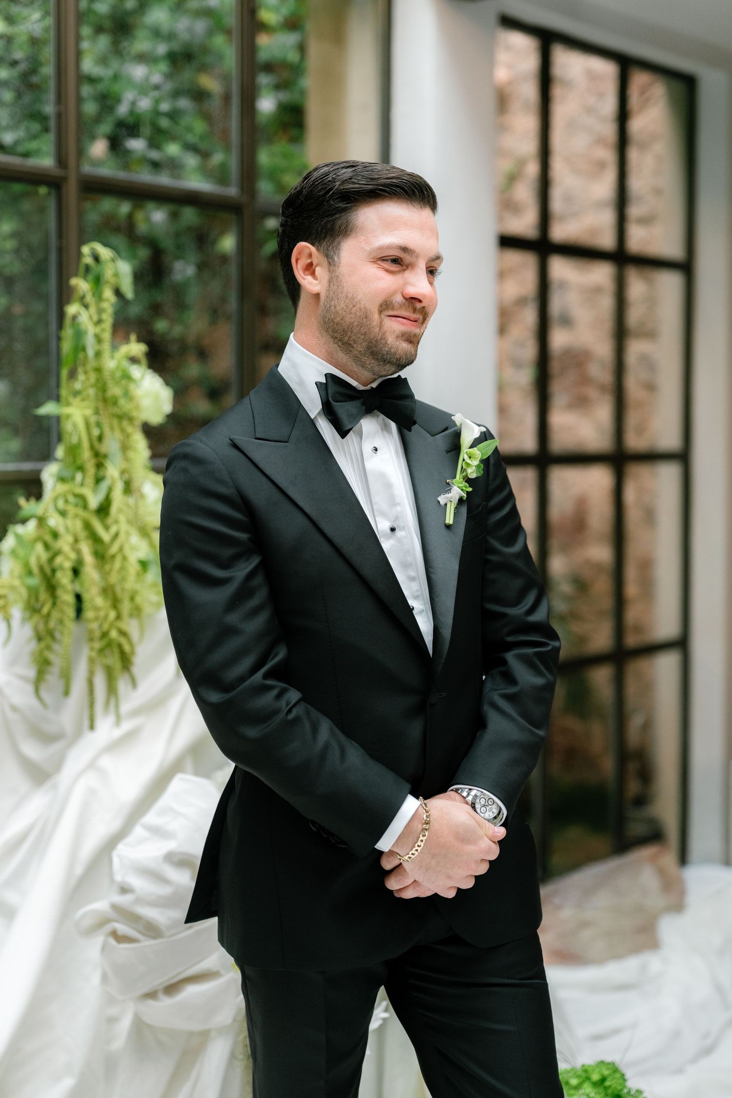 Groomsmen in black tuxedos standing in greenhouse venue with white floral decorations.