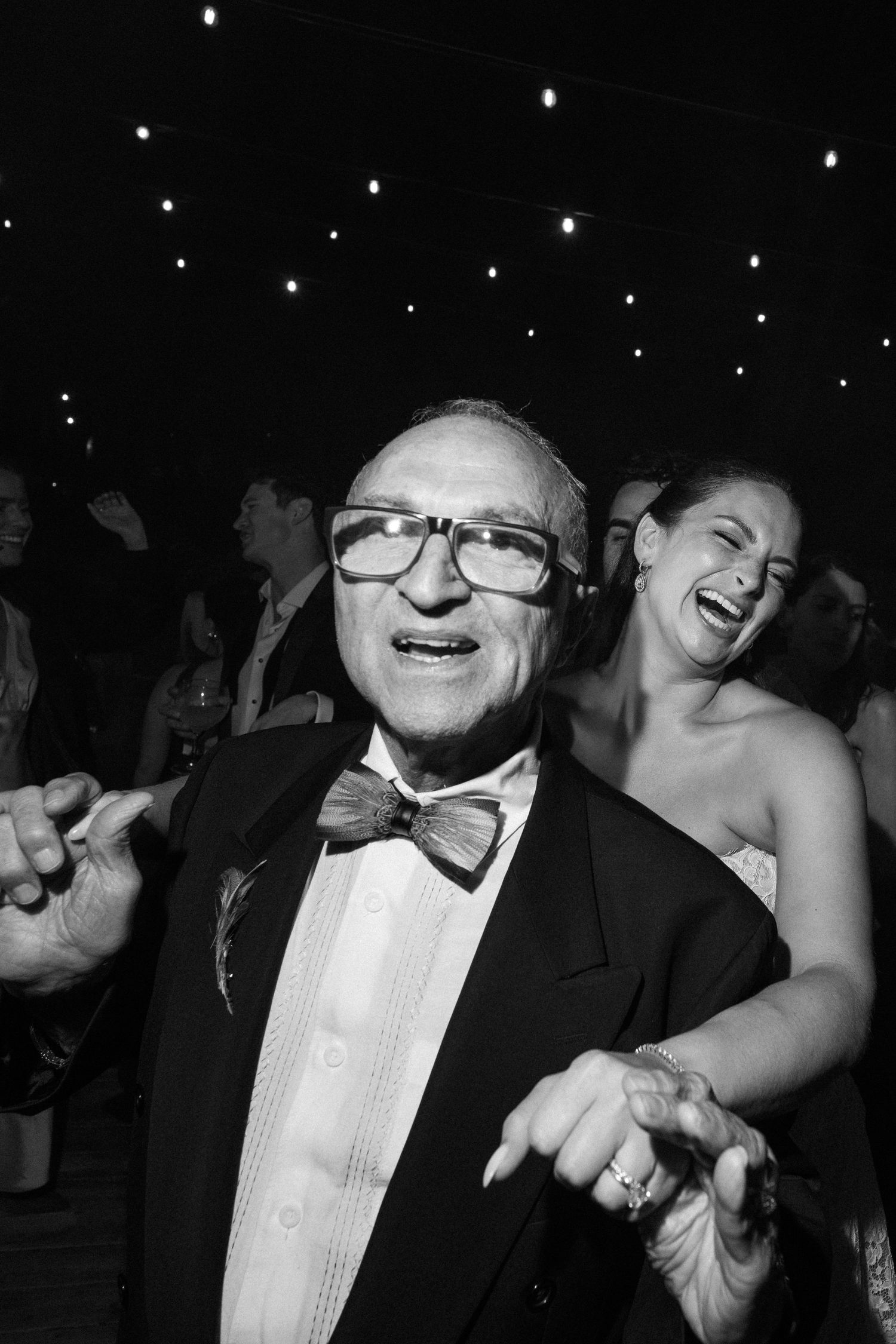 Elderly man in bow tie and glasses dancing joyfully at a wedding reception, black and white photo.