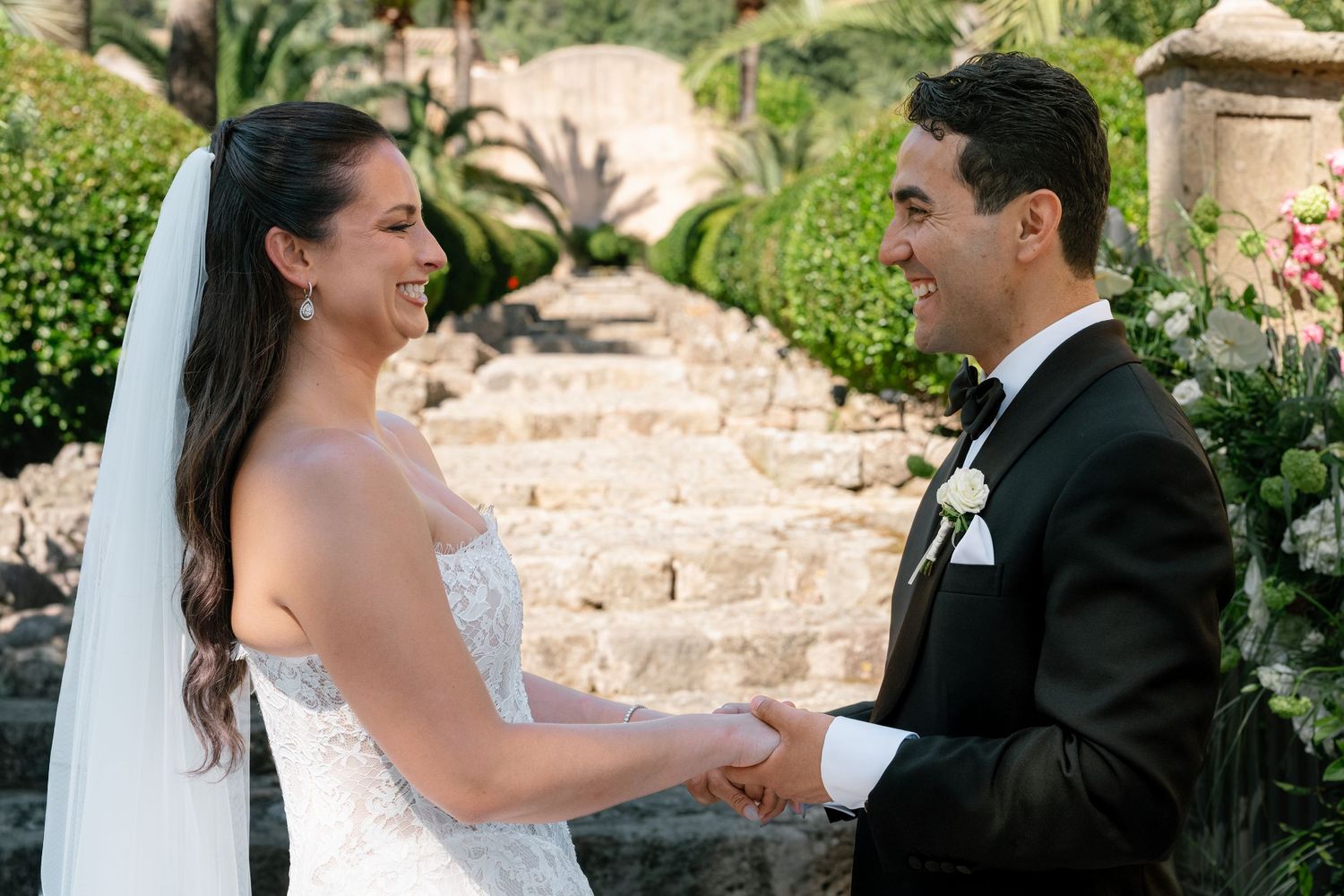 Bride and groom holding hands and smiling during outdoor wedding ceremony at elegant stone garden venue.