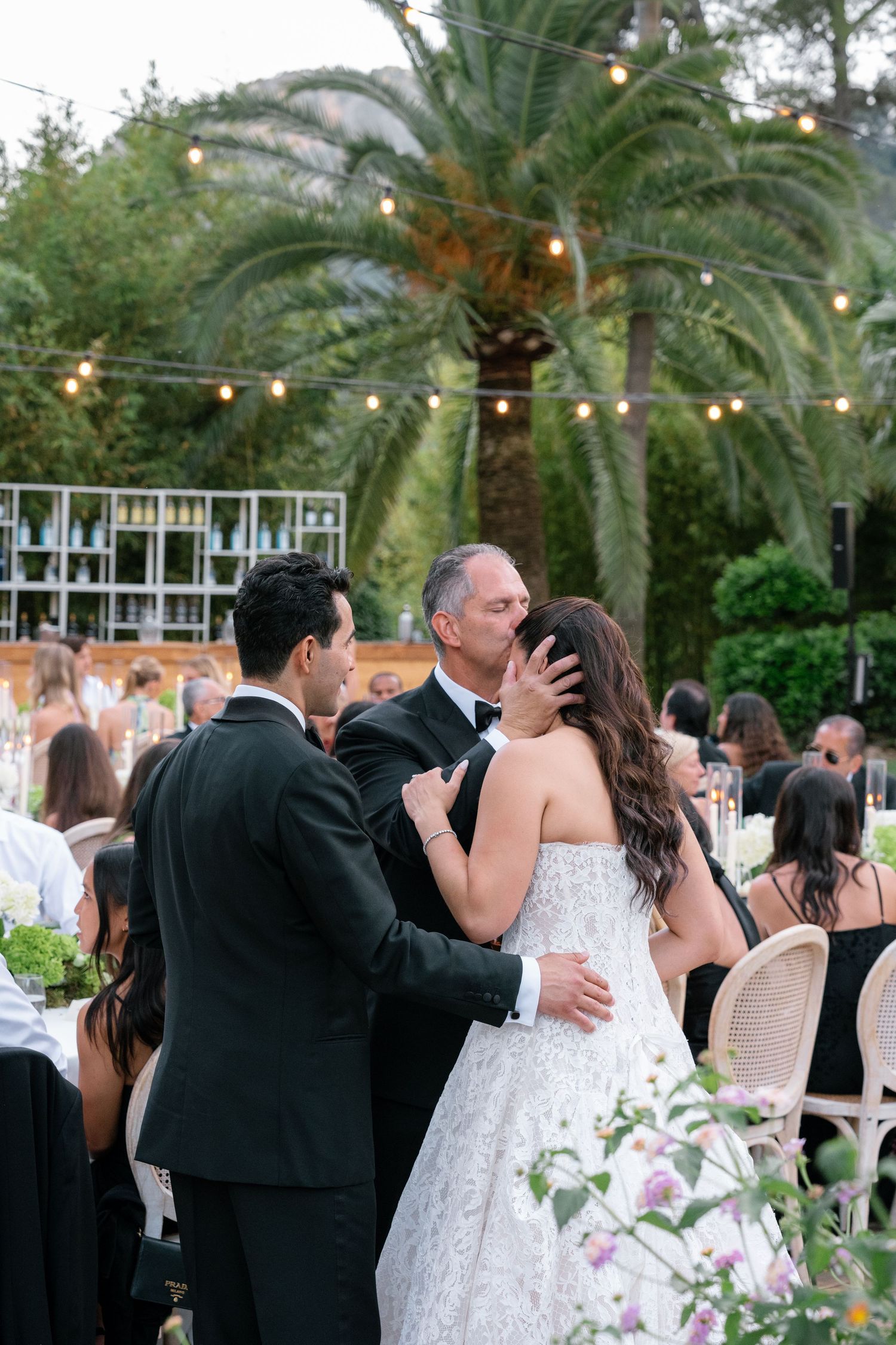 Bride in lace gown shares tender moment with groom and father during outdoor wedding reception under string lights.