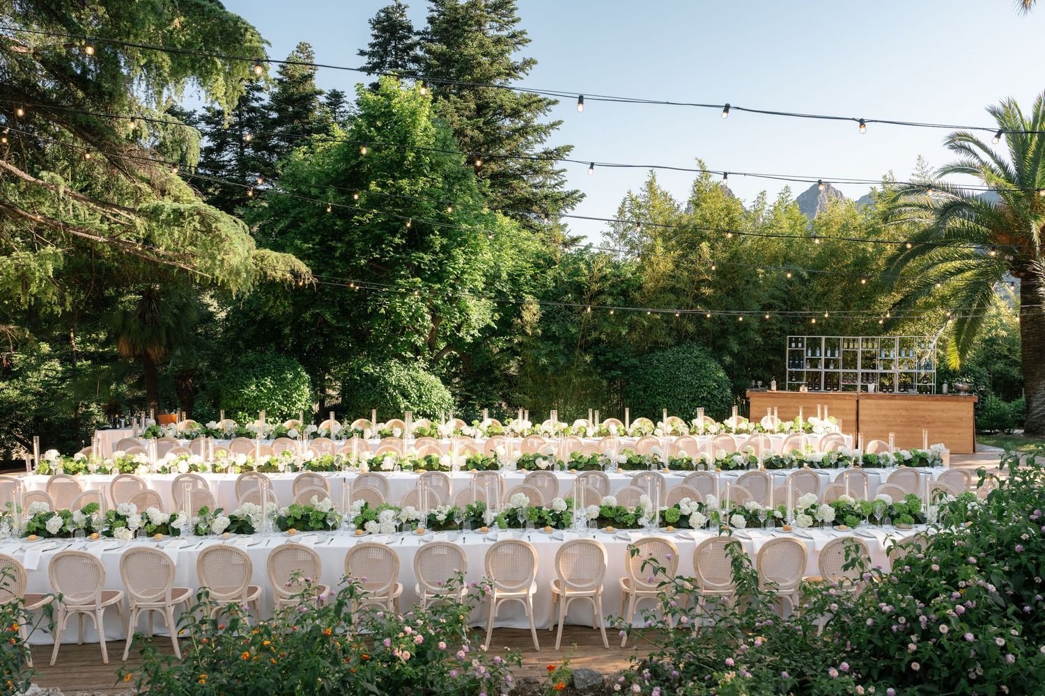 Elegant outdoor wedding reception with long tables, white chairs, floral centerpieces, string lights, and lush greenery.