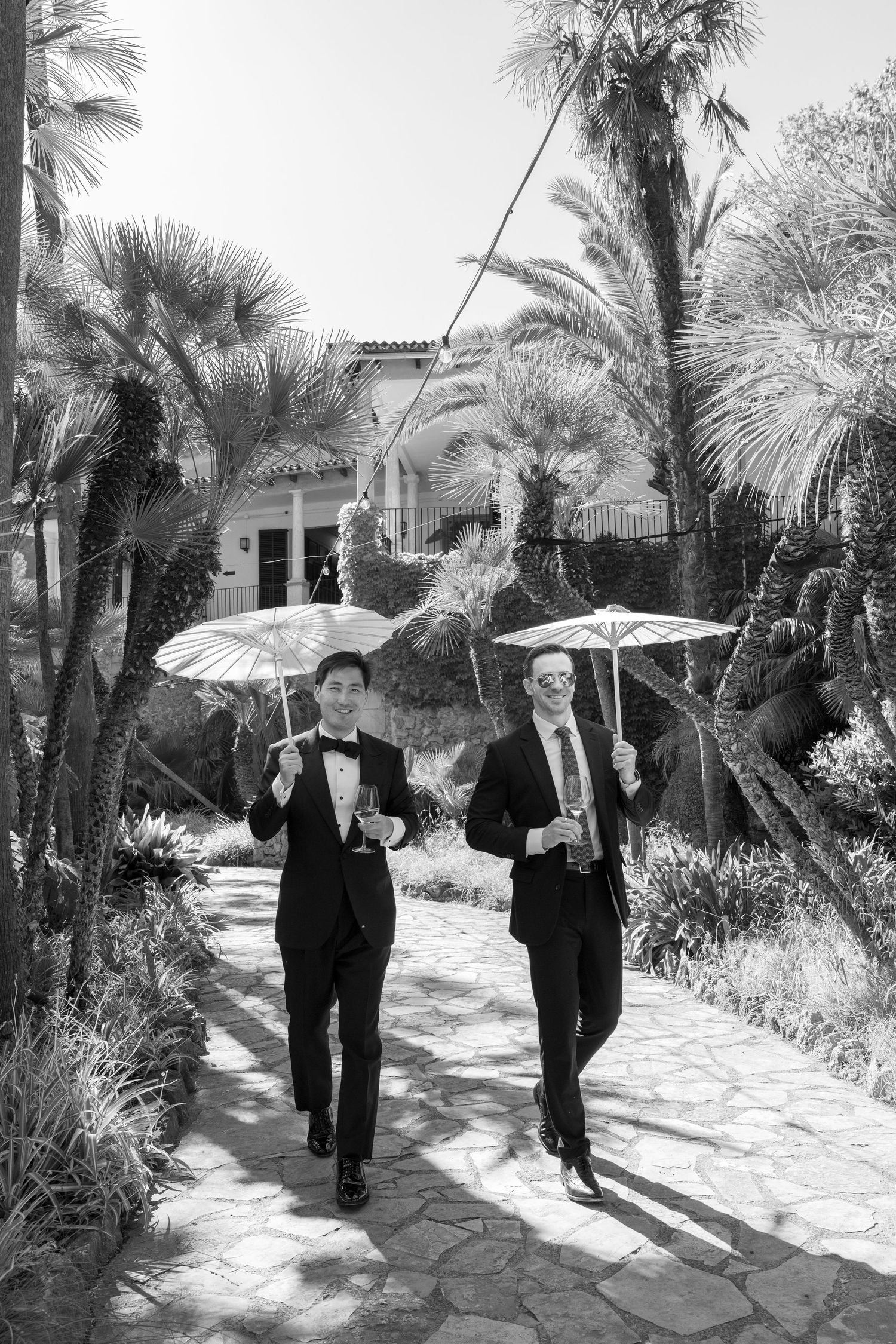 Two grooms in tuxedos walk with parasols along a stone path surrounded by palm trees at an outdoor wedding.