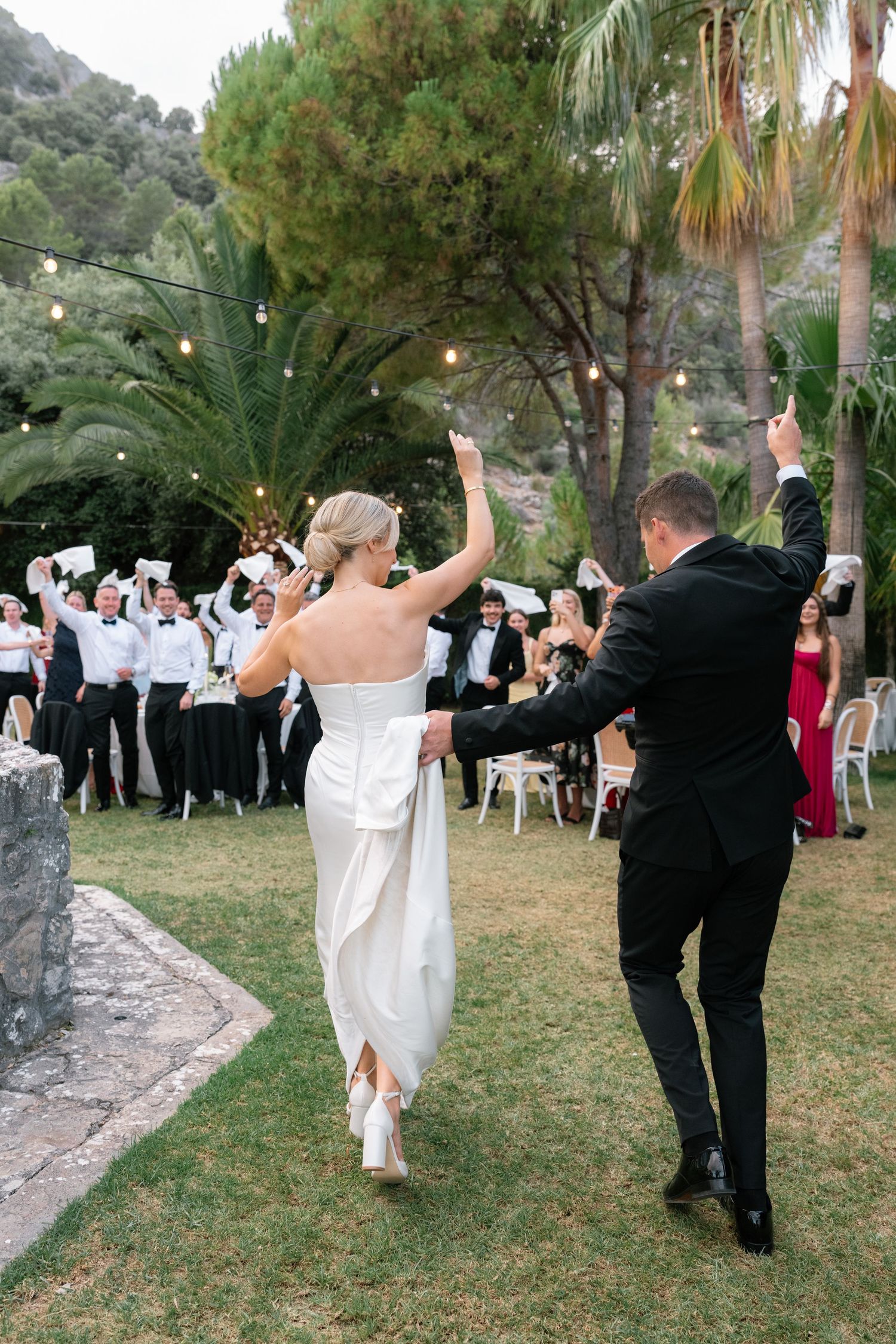 Bride and groom walk hand-in-hand toward cheering guests waving white napkins at an outdoor wedding reception.