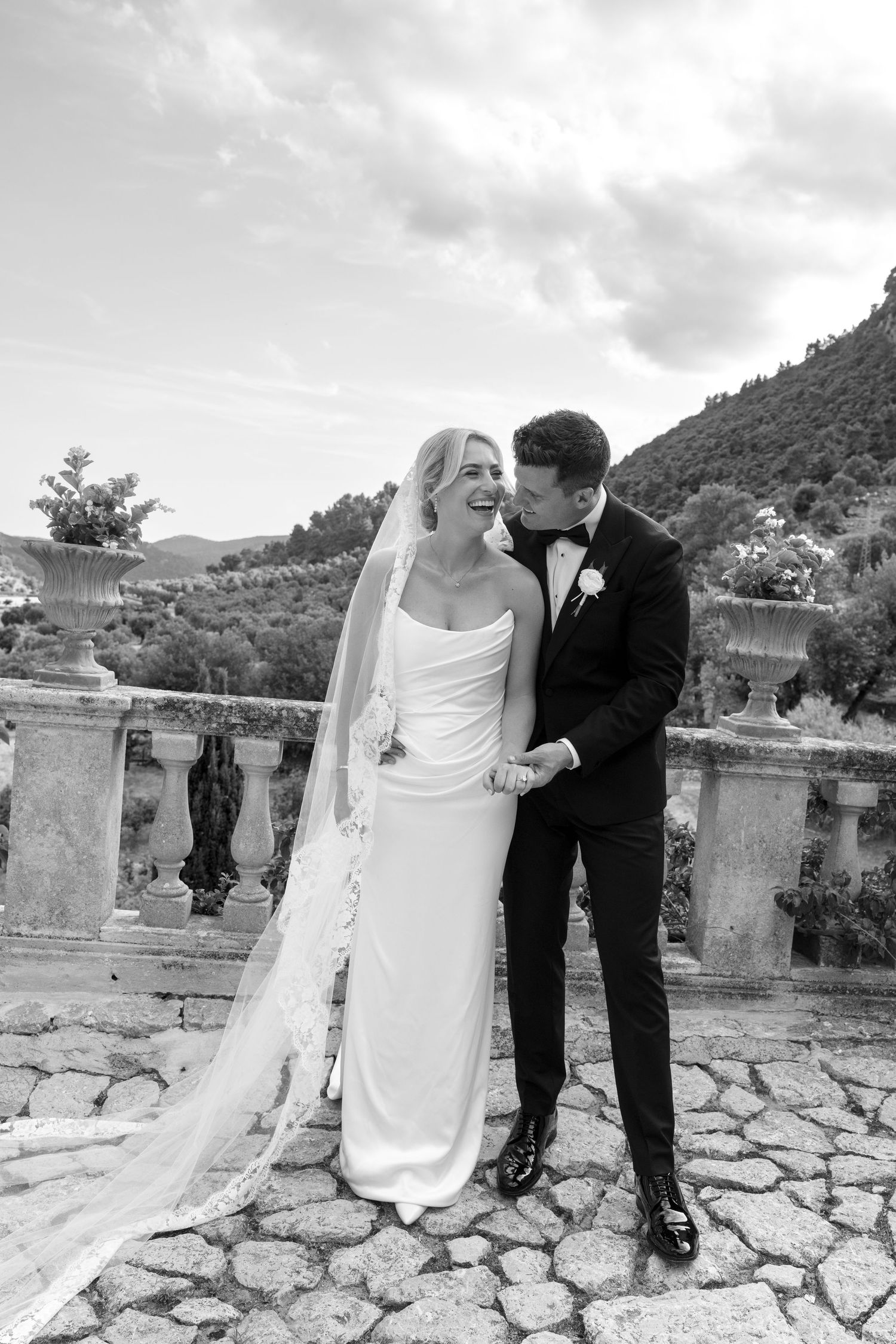 Black and white photo of a bride in a white gown and veil laughing as groom in tuxedo kisses her cheek outdoors.