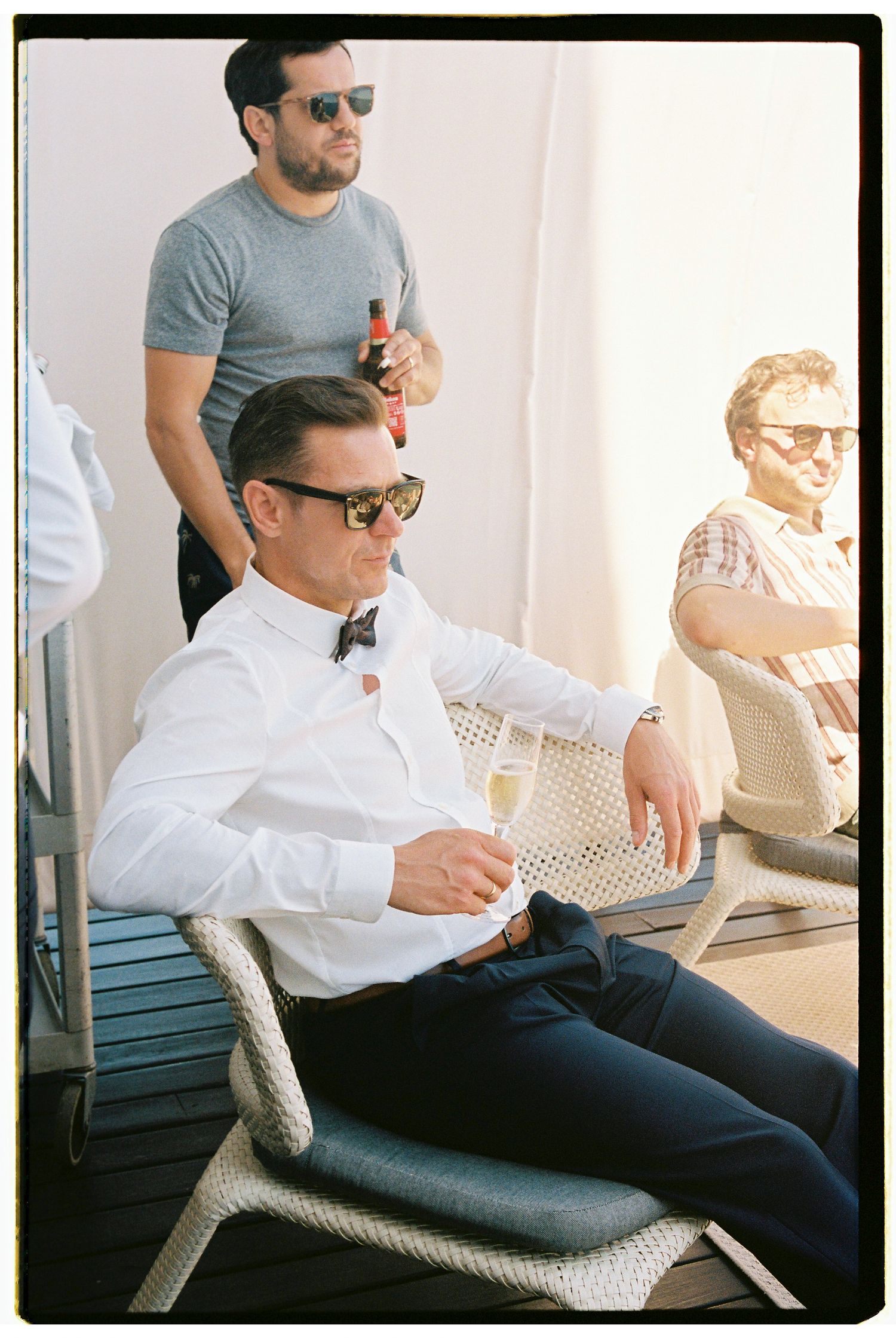 Person in sunglasses and white shirt relaxes in wicker chair while enjoying a beverage on sunny patio.
