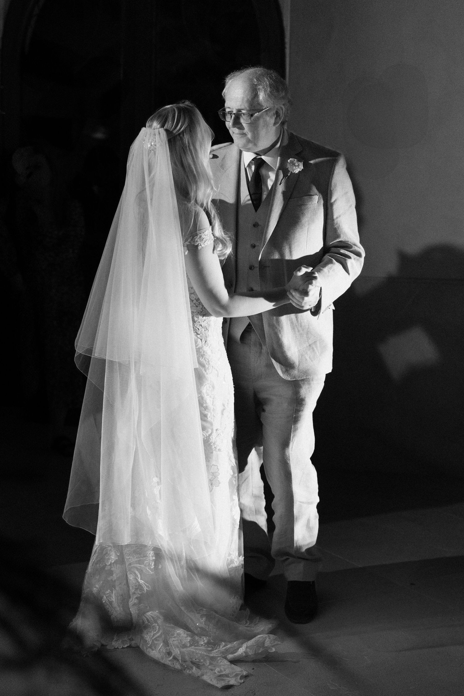 Black and white photo of a bride in a lace gown sharing a tender moment with an older man during a wedding.