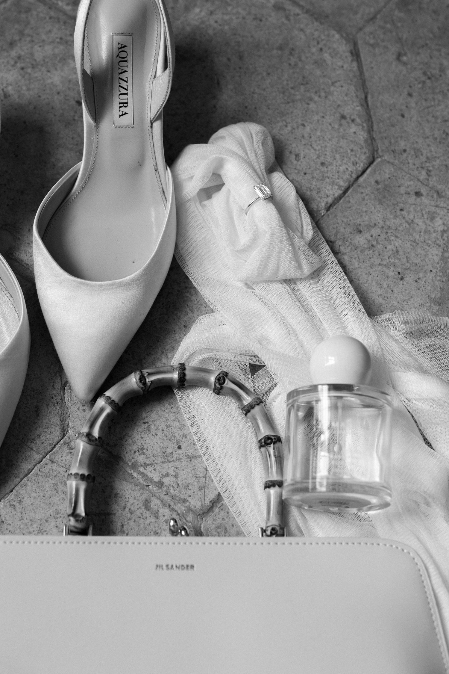 Wedding accessories including white heels, bracelet, and perfume arranged on grey surface.