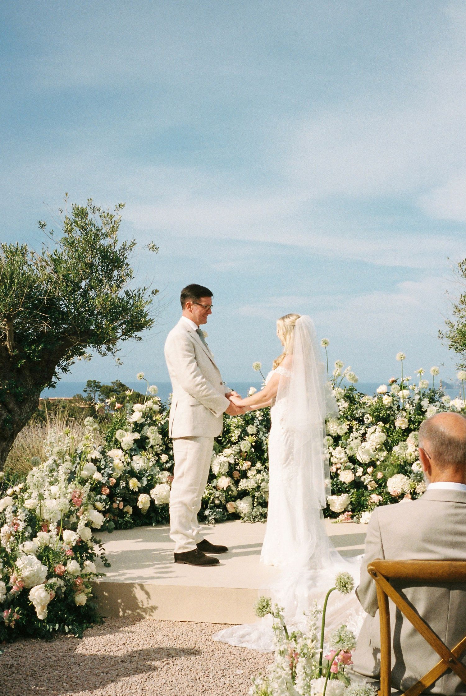 Wedding ceremony setup with white floral arrangements and wooden chairs against blue sky.