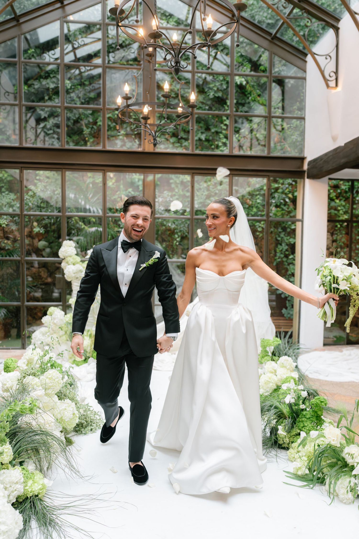 A couple in formal wedding attire dances and poses playfully in a sunlit greenhouse with white decor.