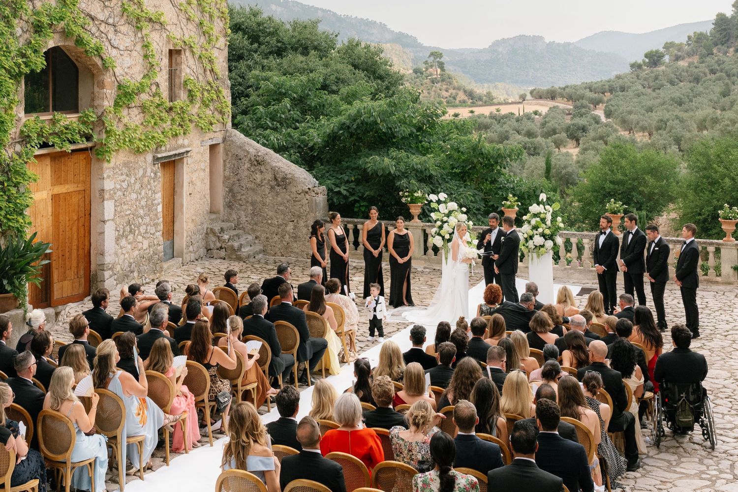 Outdoor wedding ceremony at a rustic stone villa with lush mountain views, guests seated in rows facing the altar.