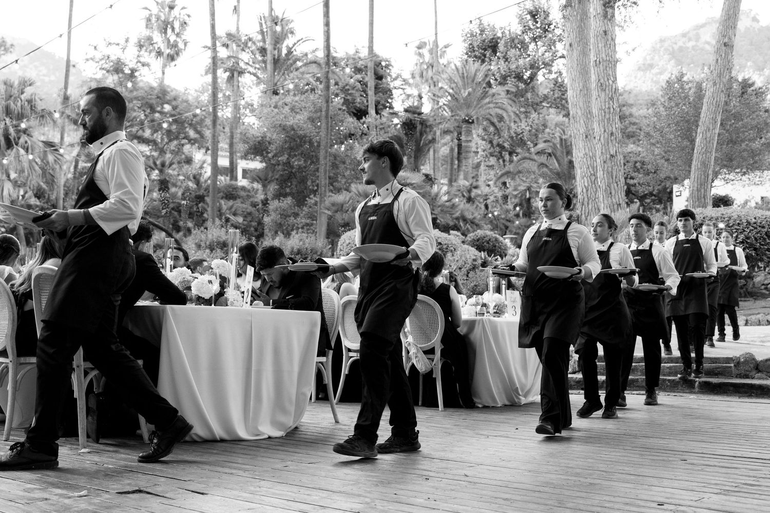 Waitstaff in black aprons carry plates at an outdoor wedding reception surrounded by lush trees.