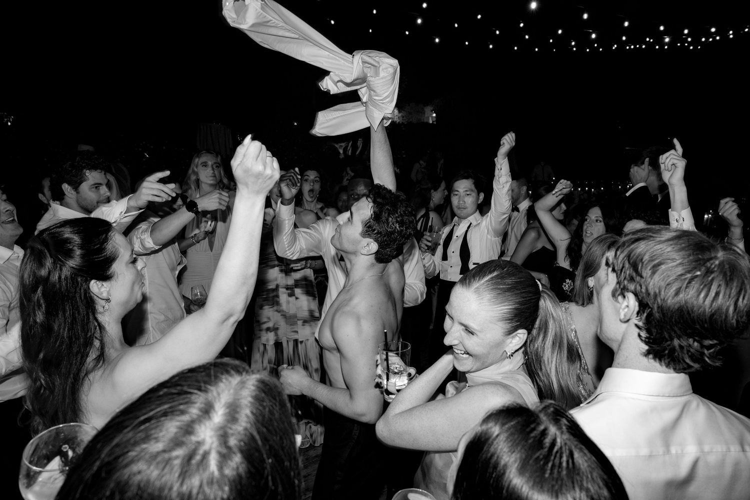 Black and white photo of a lively wedding reception dance floor with guests celebrating and raising their hands in joy.