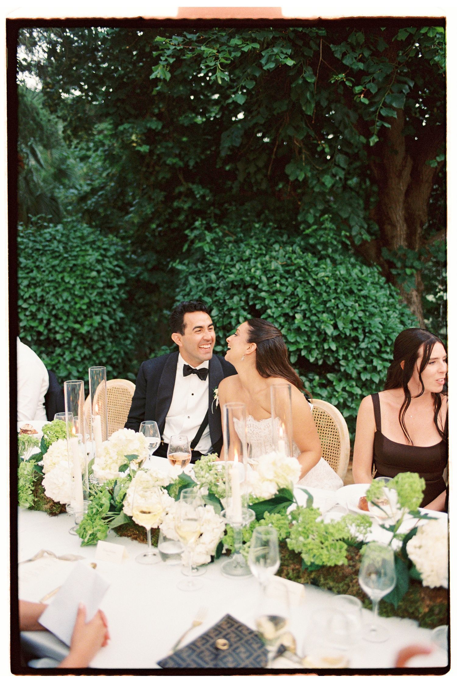 Bride and groom laughing at outdoor wedding reception table with white floral arrangements and lush green backdrop.