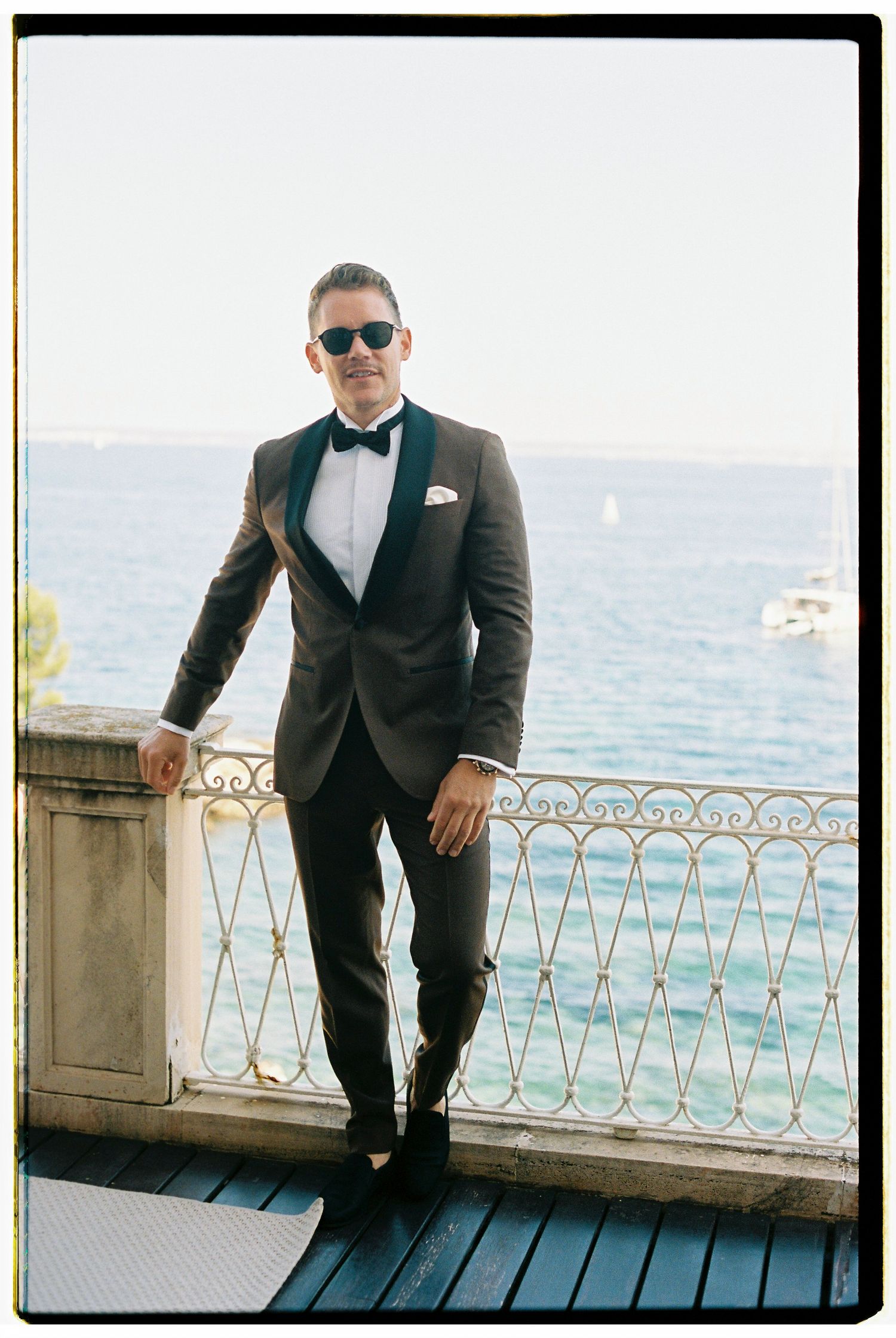 A person in a black tuxedo poses elegantly against an ornate railing overlooking the Mediterranean Sea.