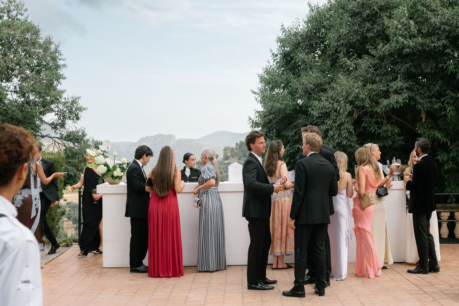 Elegantly dressed wedding guests mingle outdoors on a terrace with mountain views and lush greenery.