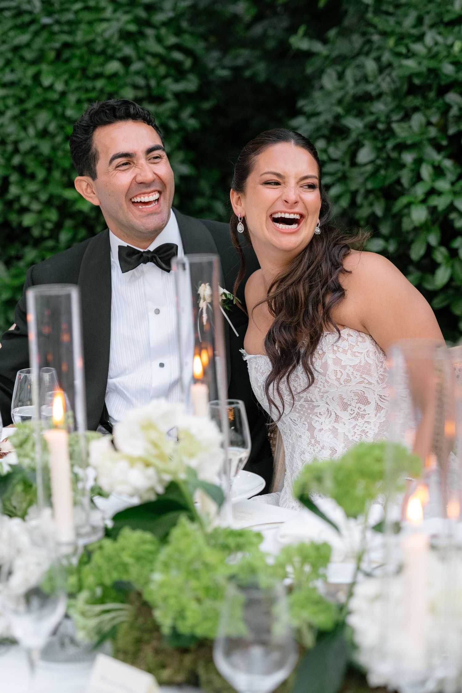 Bride and groom laughing joyfully at wedding reception table with white floral centerpieces and candles.