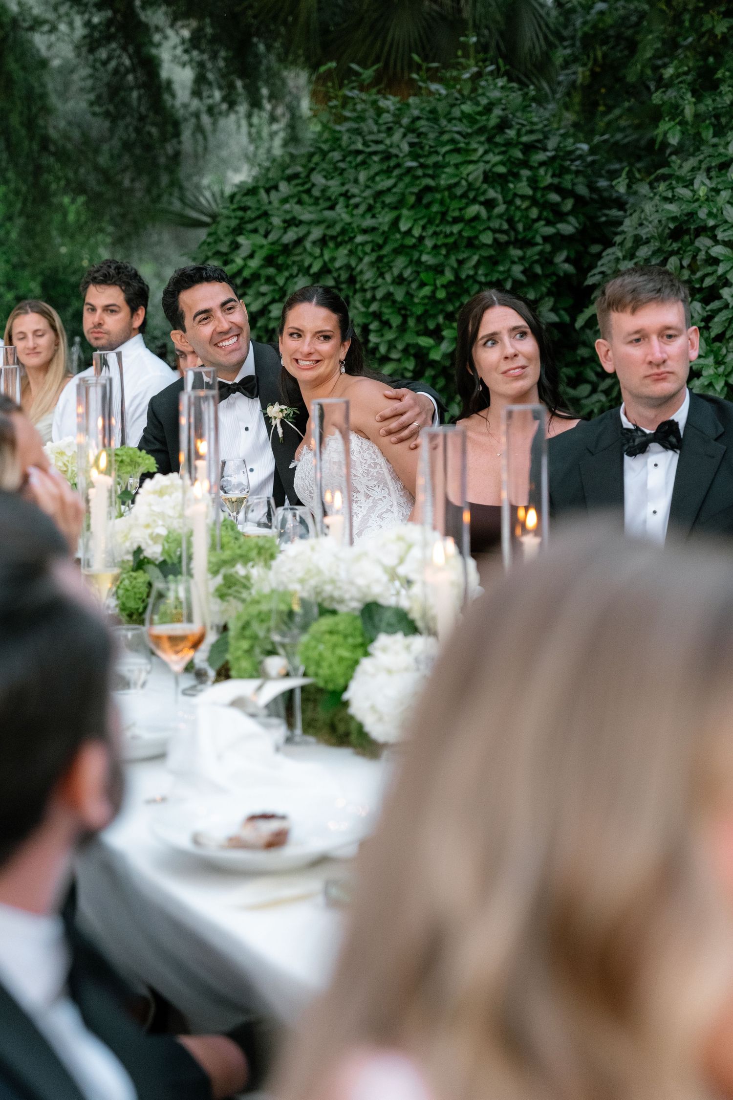 Bride and groom smile at wedding reception table adorned with white hydrangeas and greenery backdrop, guests in formal attire.