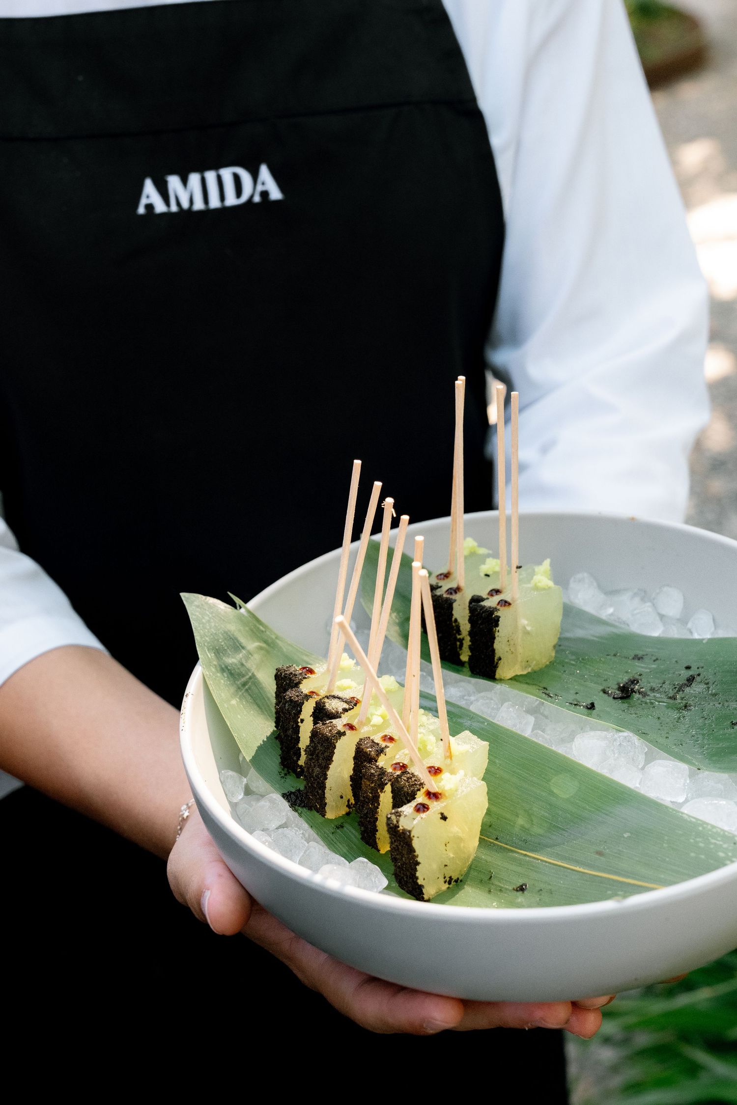 Amida restaurant server holding a white bowl with elegant skewered appetizers on banana leaf and ice.