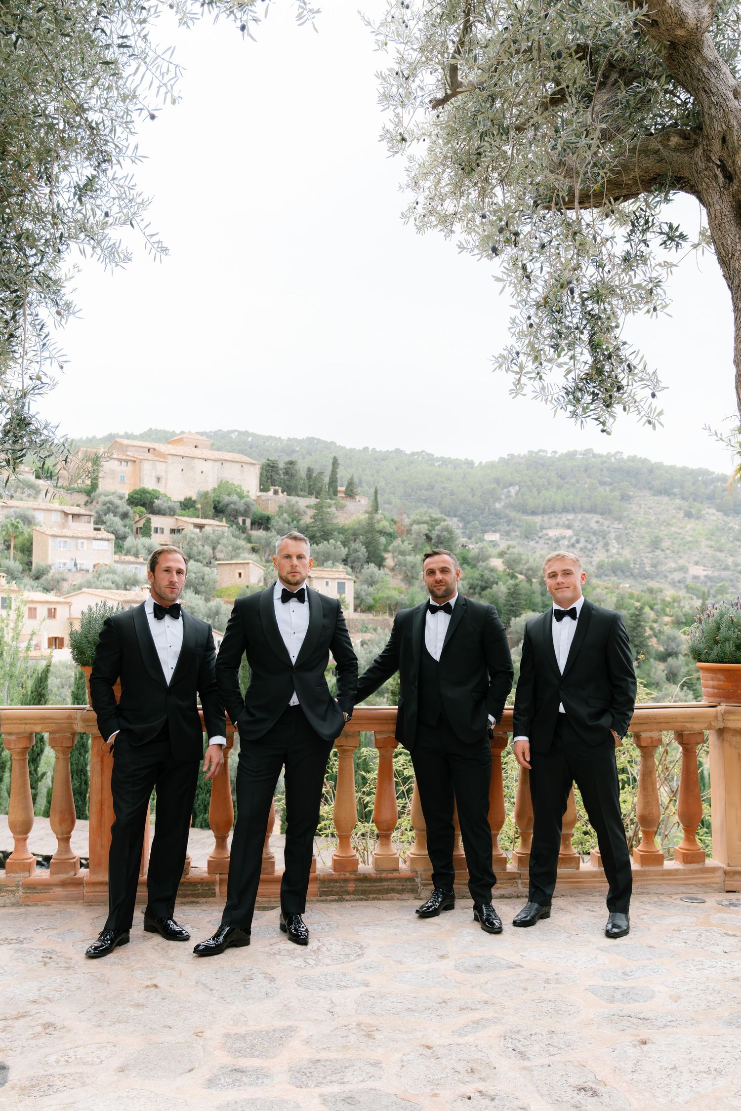 Groomsmen in black tuxedos lined up on stone balcony with Mediterranean landscape backdrop.