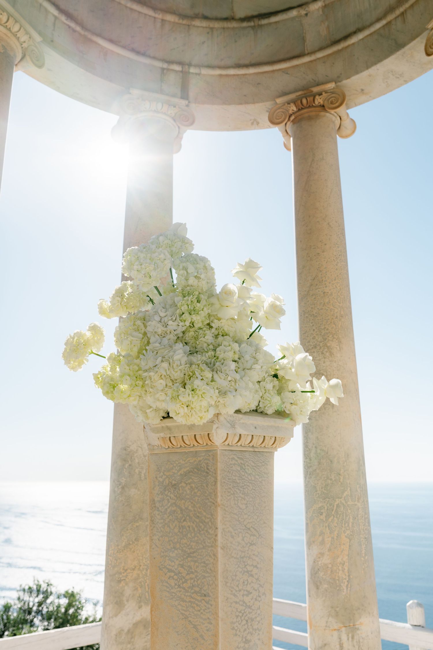 White floral arrangements decorating classical stone columns with ocean view in background.