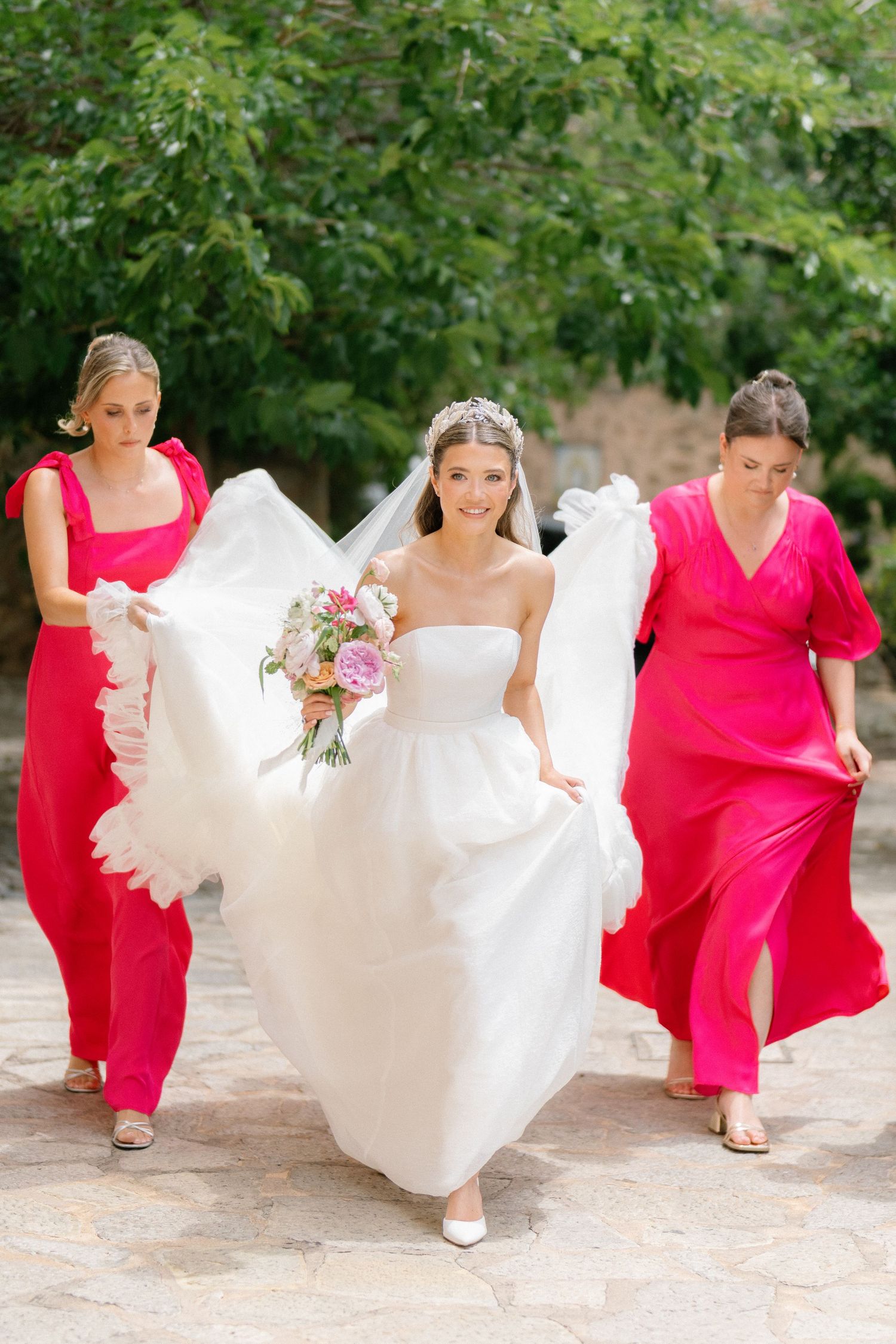 Bride in white gown walks with bridesmaids in bright pink dresses along a garden path.
