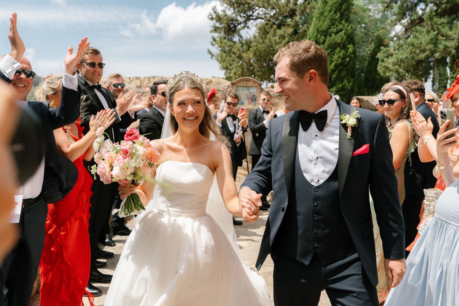 Joyful outdoor wedding celebration with guests cheering under bright blue skies.