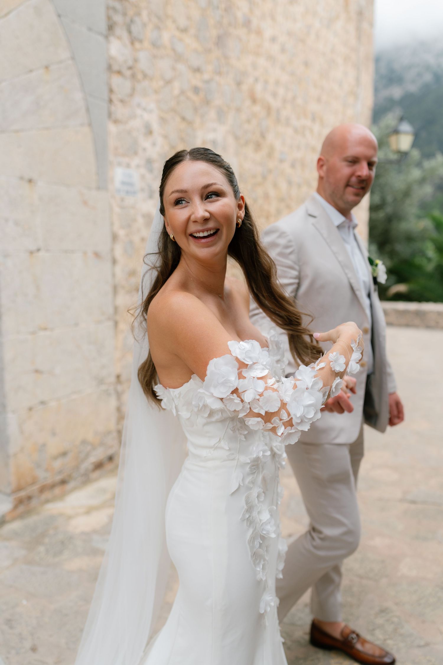 Bride in lace wedding dress with floral appliques poses against stone wall at Mediterranean venue.
