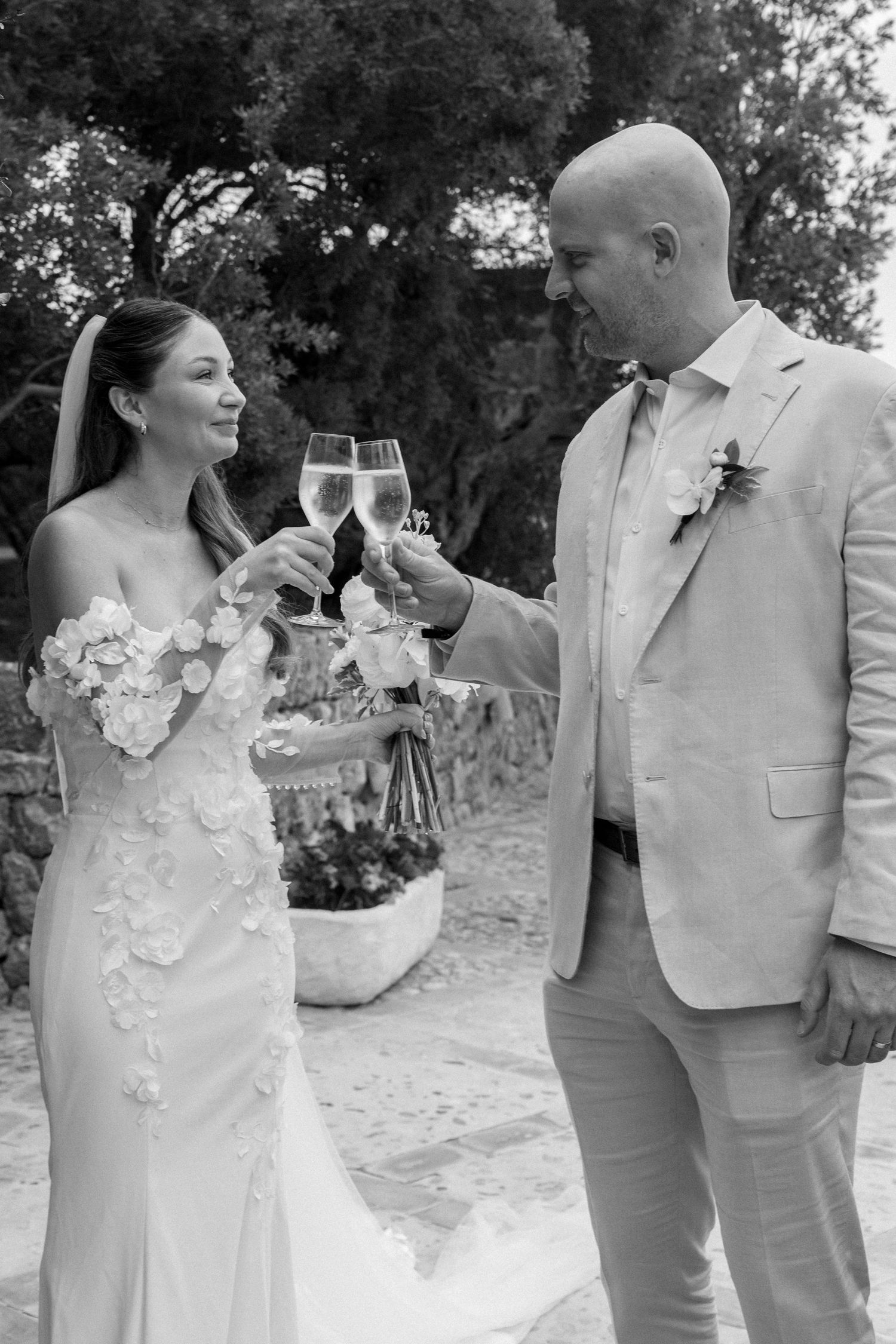 Wedding couple in light colored attire sharing champagne on an outdoor terrace.