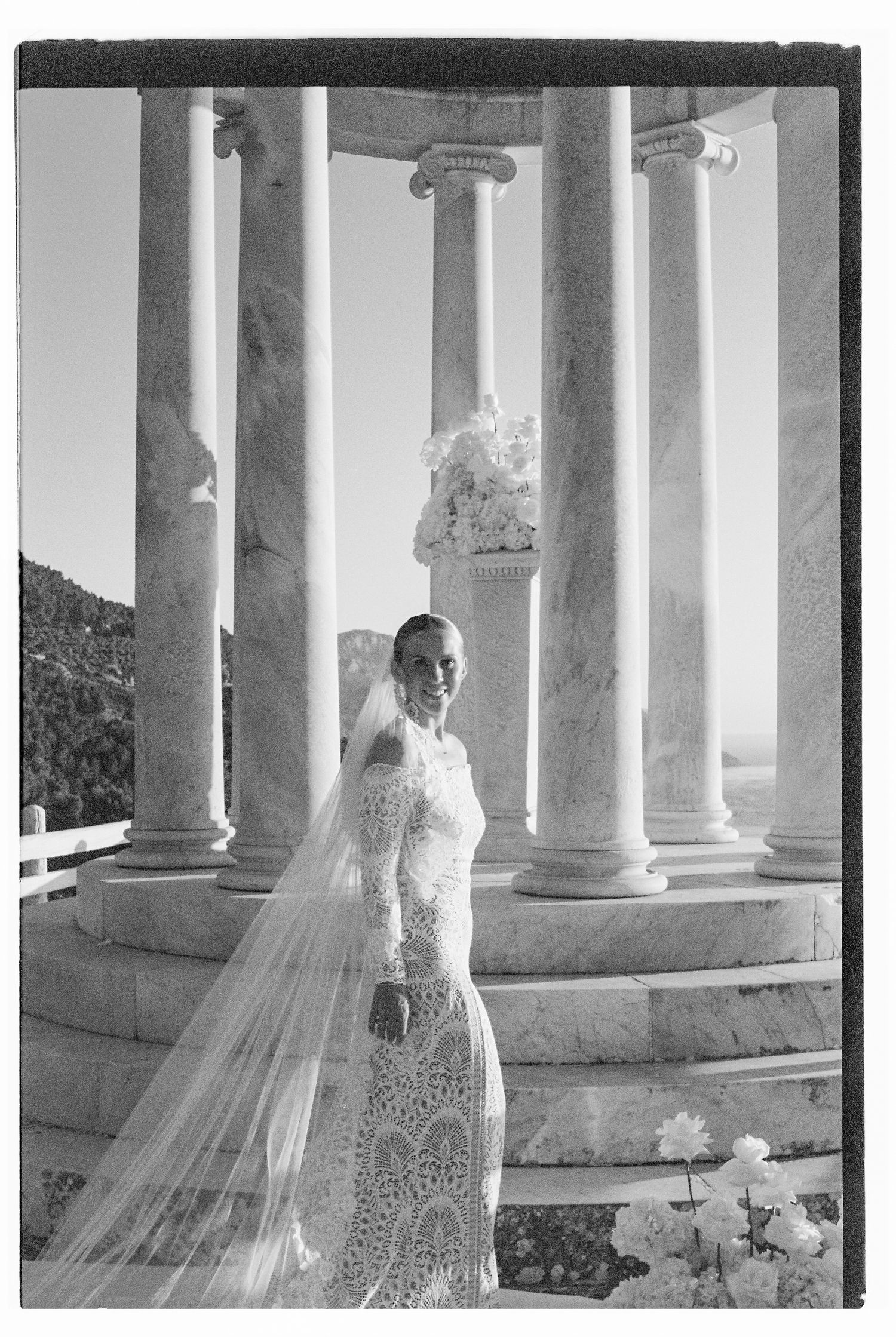 Bride in detailed lace gown standing among white marble columns with flowing veil.