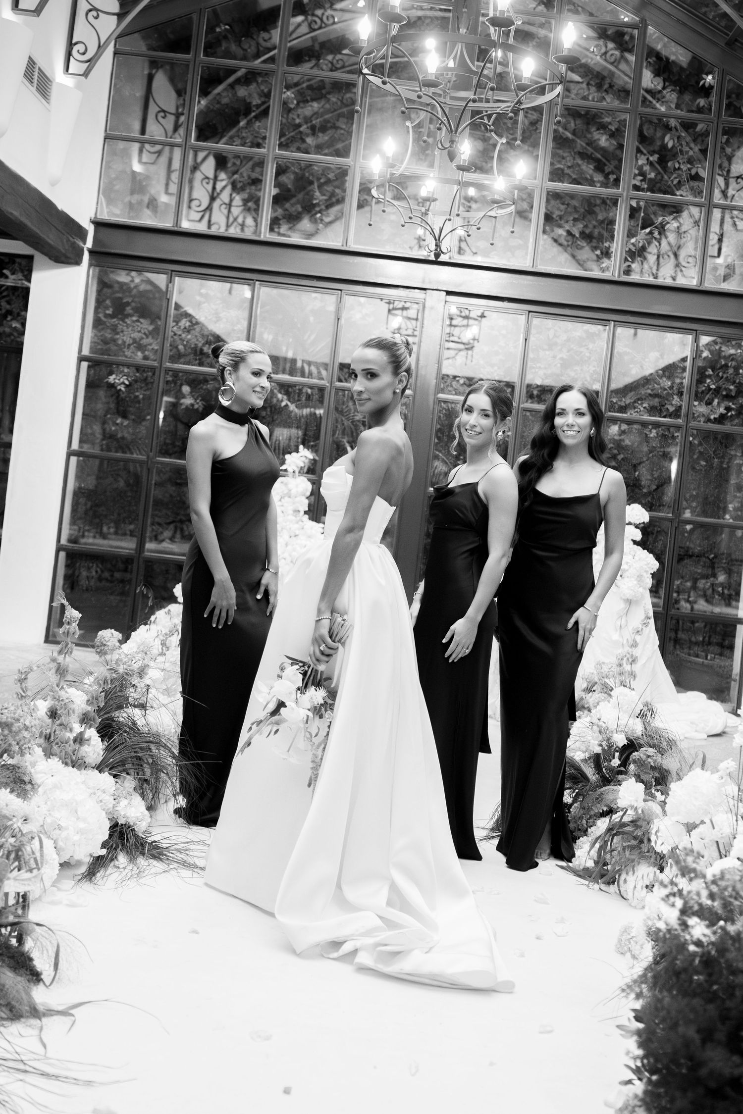 Wedding party in black and white attire poses in front of a glass conservatory with floral arrangements.