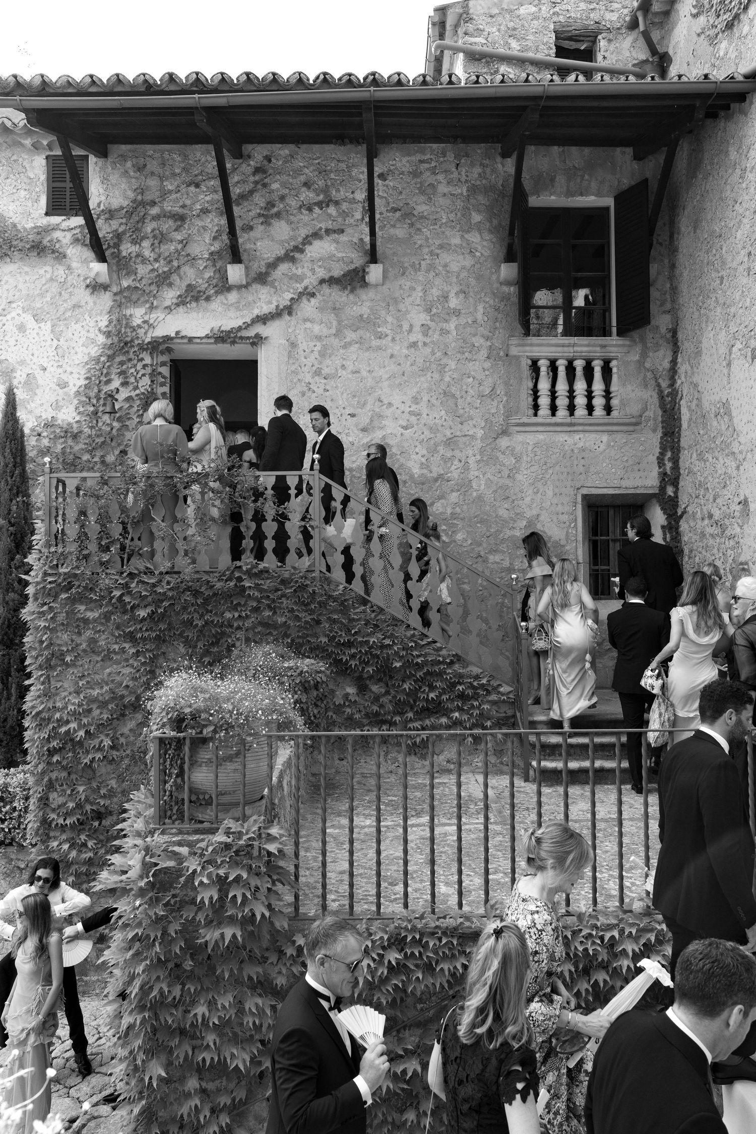 Black and white photo of elegantly dressed wedding guests gathered outside a rustic stone villa with ivy-covered walls.