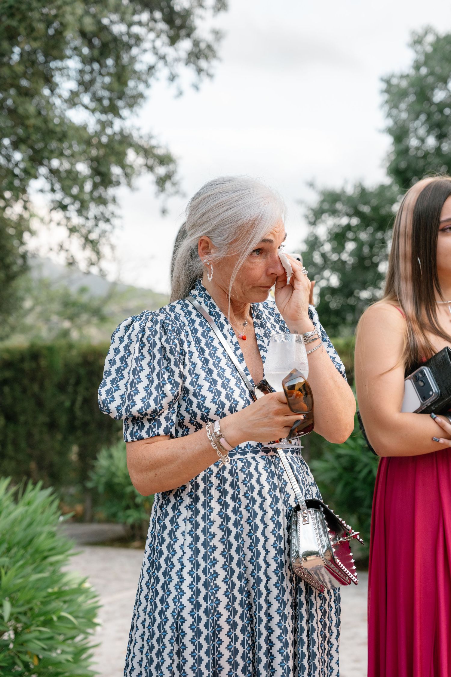 Elderly woman with gray hair wiping tears in blue patterned dress at outdoor wedding event.