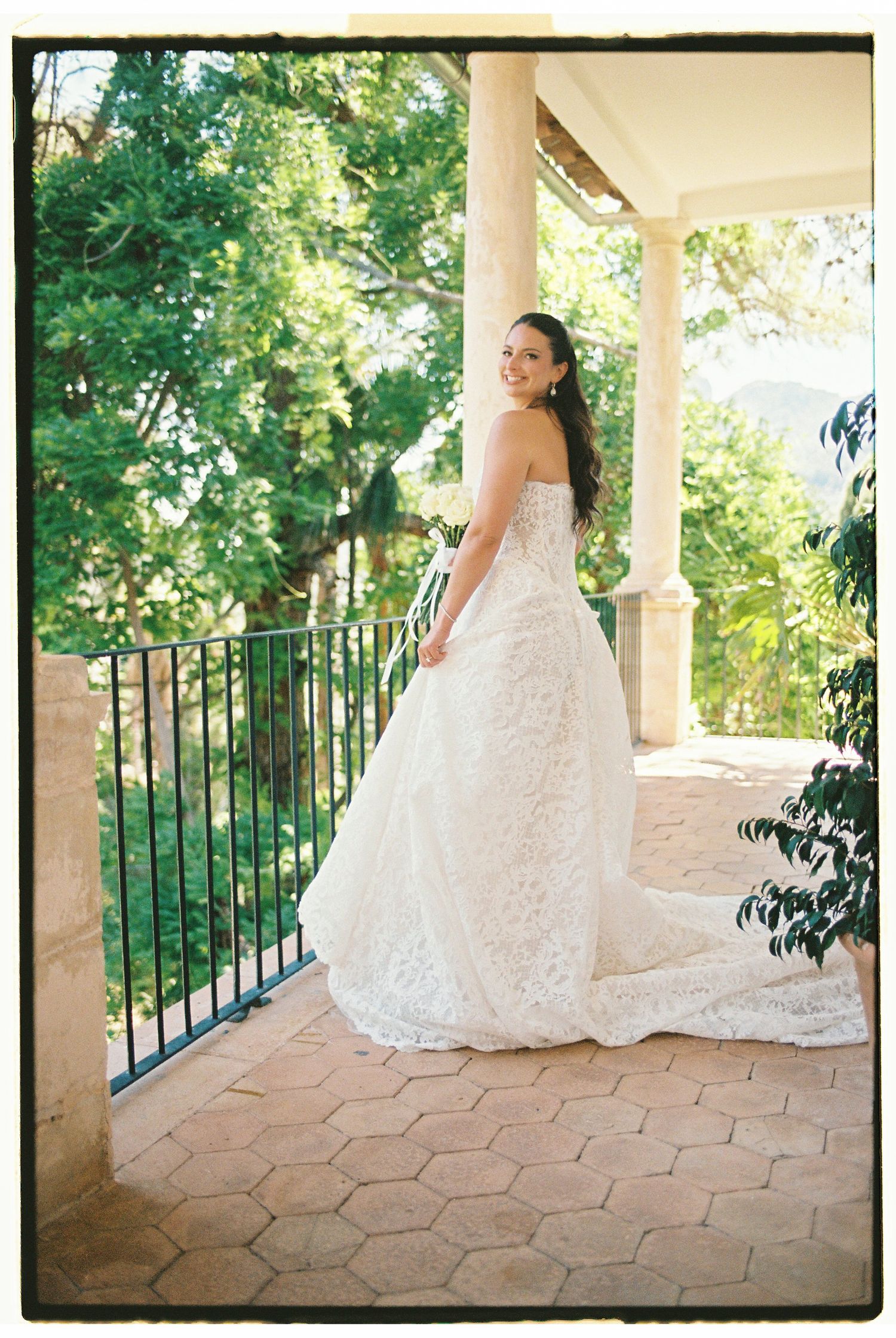 Bride in a strapless lace wedding gown with a train poses on a sunlit balcony surrounded by lush green trees.